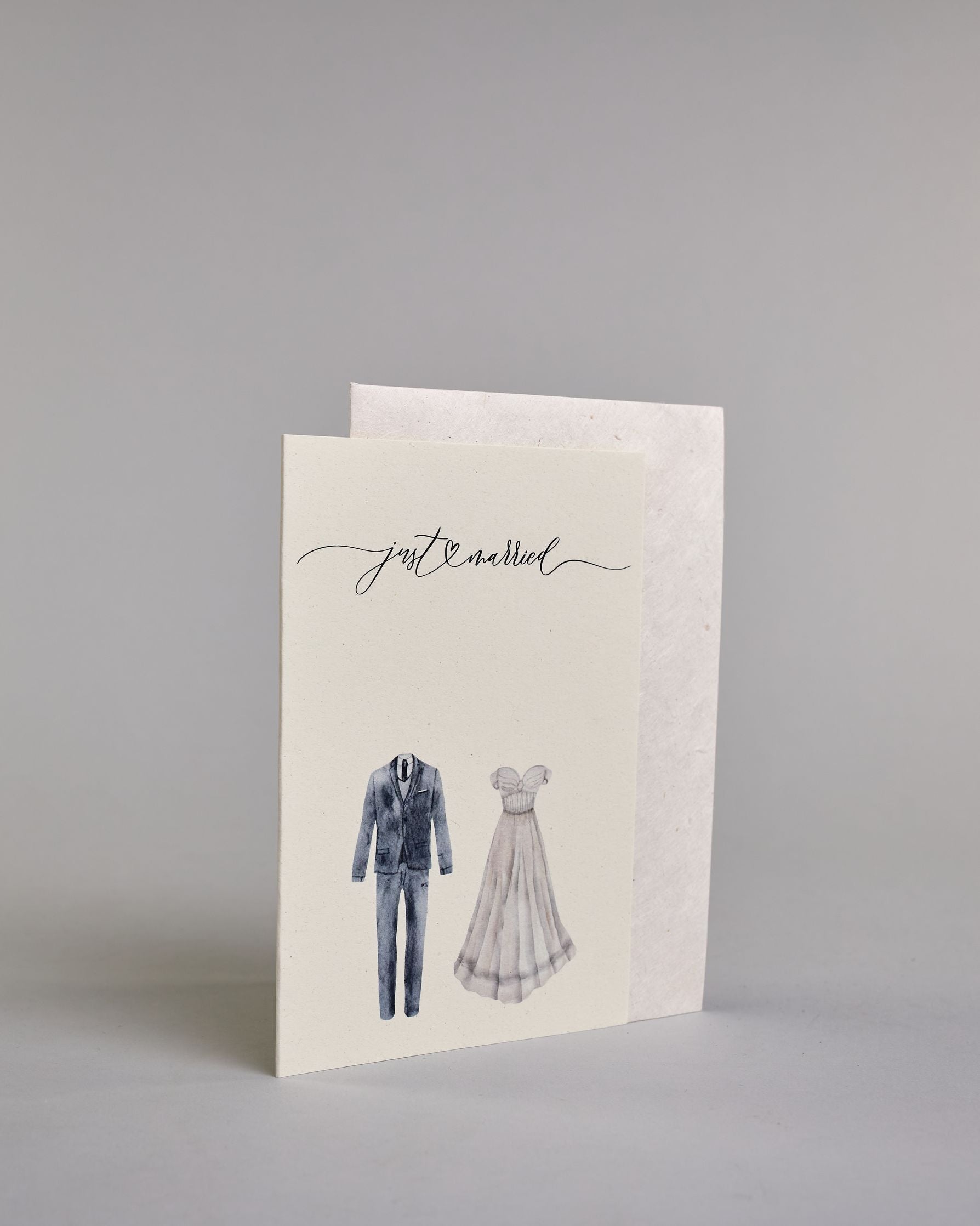 Glückwunschkarte auf Englisch zur Hochzeit mit schwarzer Kalligrafieaufschrift "just married", darunter ein schwarzer Anzug und rechts daneben ein weißes Hochzeitskleid. Der Text "just married" ist in geschwungener schwarzer Kalligraphieschrift und zieht sich von ganz links nach ganz rechts über die Karte, außerdem befindet sich im Schwung der Schrift in der Mitte der beiden Worte ein Herz und alles ist auf hellem Hintergrund. Dazu ein weißes handgeschöpftes Kuvert. #3