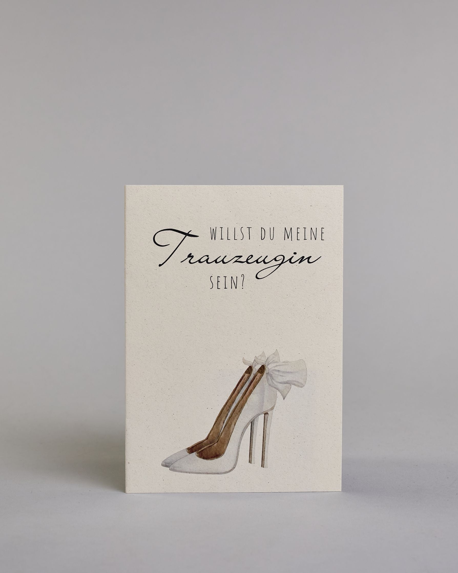Klappkarte mit der Aufschrift "Willst du meine Trauzeugin sein?" in schwarzer Schrift auf hellem Hintergrund. Darunter ist ein eleganter weißer High Heel mit einer großen Schleife an der Ferse abgebildet. Der Text ist in drei Zeilen verfasst. "WILLST DU" ist in dünner schwarzer Kalligraphieschrift über dem Wort "Trauzeugin", welches in großer geschwungener Schreibschrift gestaltet ist, dargestellt. Darunter ist in ebenfalls dünner Kalligrafieschrift "SEIN?". Alles ist auf hellem Hintergrund und dazu kommt…