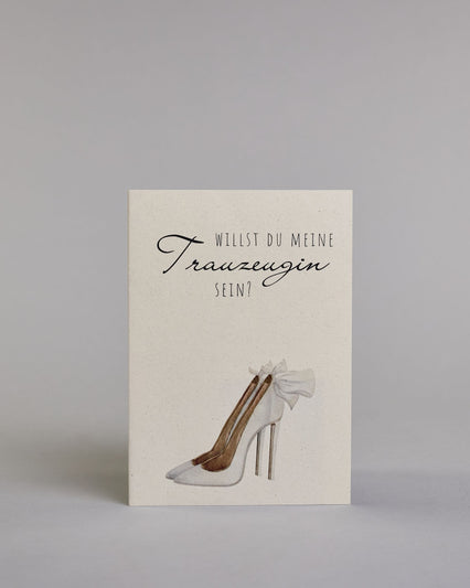 Klappkarte mit der Aufschrift "Willst du meine Trauzeugin sein?" in schwarzer Schrift auf hellem Hintergrund. Darunter ist ein eleganter weißer High Heel mit einer großen Schleife an der Ferse abgebildet. Der Text ist in drei Zeilen verfasst. "WILLST DU" ist in dünner schwarzer Kalligraphieschrift über dem Wort "Trauzeugin", welches in großer geschwungener Schreibschrift gestaltet ist, dargestellt. Darunter ist in ebenfalls dünner Kalligrafieschrift "SEIN?". Alles ist auf hellem Hintergrund und dazu kommt…