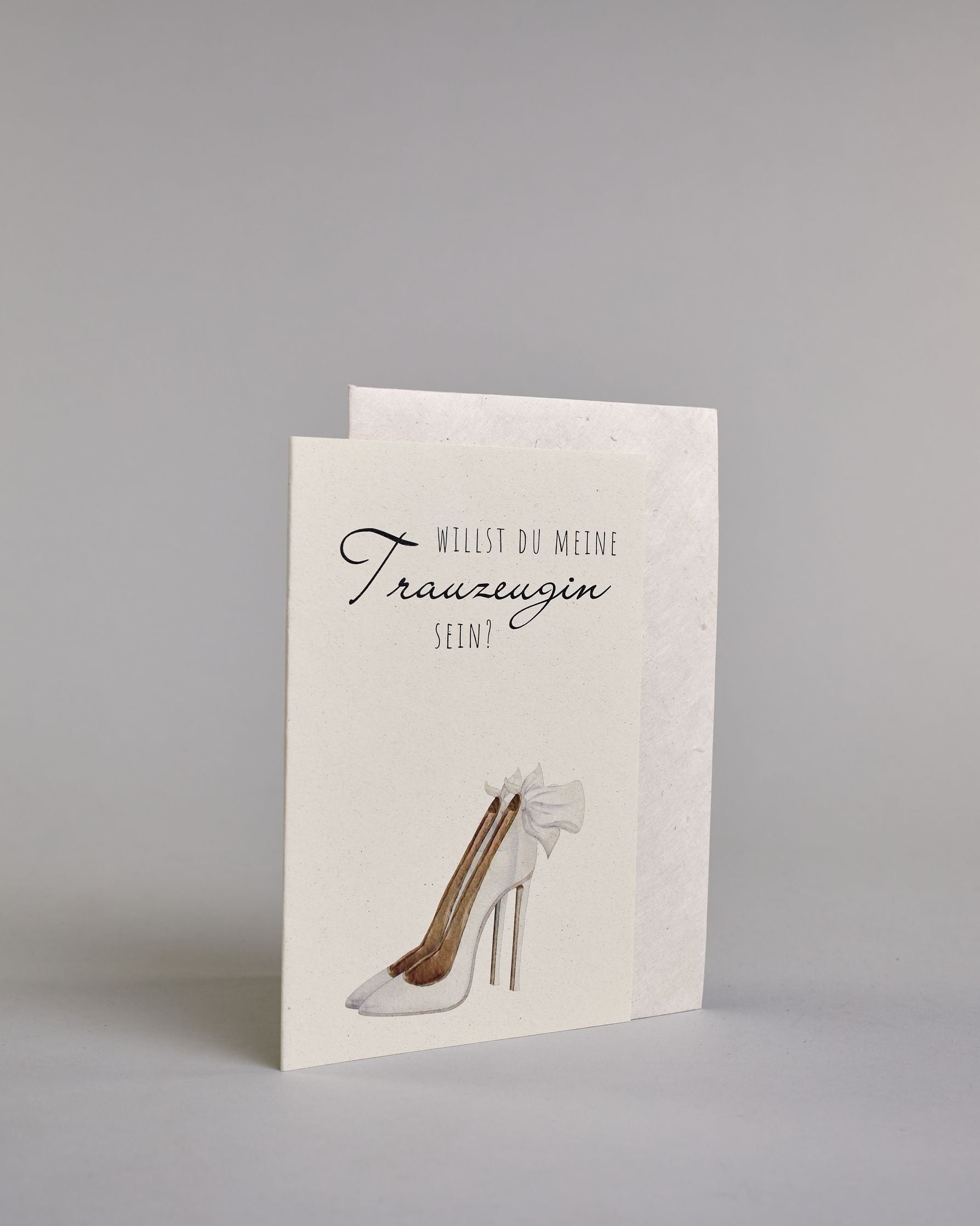 Klappkarte mit der Aufschrift "Willst du meine Trauzeugin sein?" in schwarzer Schrift auf hellem Hintergrund. Darunter ist ein eleganter weißer High Heel mit einer großen Schleife an der Ferse abgebildet. Der Text ist in drei Zeilen verfasst. "WILLST DU" ist in dünner schwarzer Kalligraphieschrift über dem Wort "Trauzeugin", welches in großer geschwungener Schreibschrift gestaltet ist, dargestellt. Darunter ist in ebenfalls dünner Kalligrafieschrift "SEIN?". Alles ist auf hellem Hintergrund und dazu kommt…
