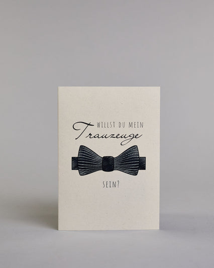 Klappkarte mit der Aufschrift "Willst du meine Trauzeugin sein?" in schwarzer Schrift auf hellem Hintergrund. Darunter ist ein eleganter weißer High Heel mit einer großen Schleife an der Ferse abgebildet. Der Text ist in drei Zeilen verfasst. "WILLST DU" ist in dünner schwarzer Kalligraphieschrift über dem Wort "Trauzeugin", welches in großer geschwungener Schreibschrift gestaltet ist, dargestellt. Darunter ist in ebenfalls dünner Kalligrafieschrift "SEIN?". Alles ist auf hellem Hintergrund und dazu kommt…