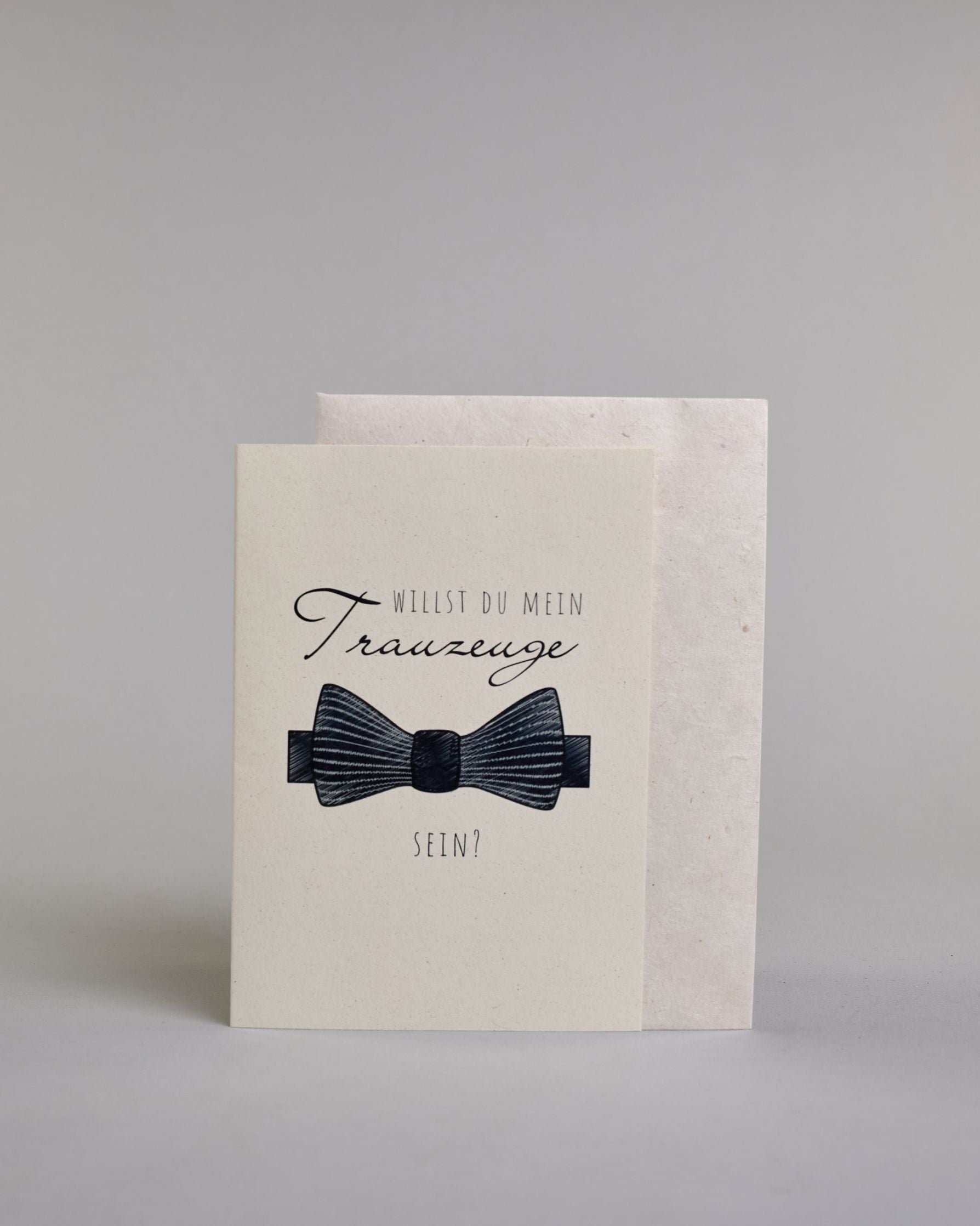 Klappkarte mit der Aufschrift "Willst du meine Trauzeugin sein?" in schwarzer Schrift auf hellem Hintergrund. Darunter ist ein eleganter weißer High Heel mit einer großen Schleife an der Ferse abgebildet. Der Text ist in drei Zeilen verfasst. "WILLST DU" ist in dünner schwarzer Kalligraphieschrift über dem Wort "Trauzeugin", welches in großer geschwungener Schreibschrift gestaltet ist, dargestellt. Darunter ist in ebenfalls dünner Kalligrafieschrift "SEIN?". Alles ist auf hellem Hintergrund und dazu kommt…
