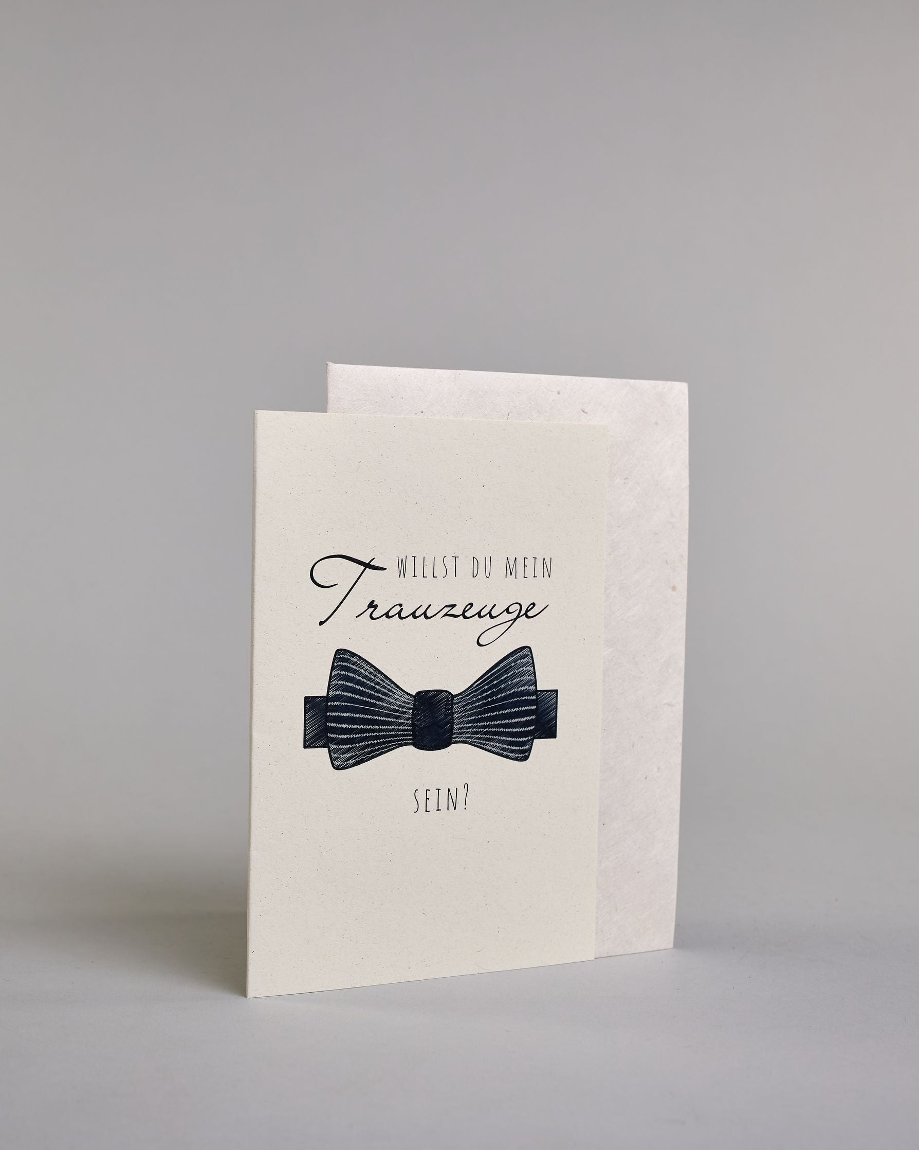 Klappkarte mit der Aufschrift "Willst du meine Trauzeugin sein?" in schwarzer Schrift auf hellem Hintergrund. Darunter ist ein eleganter weißer High Heel mit einer großen Schleife an der Ferse abgebildet. Der Text ist in drei Zeilen verfasst. "WILLST DU" ist in dünner schwarzer Kalligraphieschrift über dem Wort "Trauzeugin", welches in großer geschwungener Schreibschrift gestaltet ist, dargestellt. Darunter ist in ebenfalls dünner Kalligrafieschrift "SEIN?". Alles ist auf hellem Hintergrund und dazu kommt…
