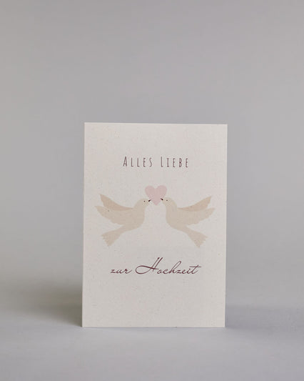 Glückwunschkarte zur Hochzeit mit bordeauxfarbener Kalligrafieaufschrift "Alles Liebe zur Hochzeit". Zwischen den Zeilen "ALLES LIEBE", welche in dünner Schrift gestaltet und der Zeile "zur Hochzeit" welche in geschwungener Schreibschrift gemacht wurde, befinden sich zwei weiße Tauben die gemeinsam mit ihren Schnäbeln ein zartrosanes Herz tragen. Alles ist auf hellem Hintergrund. Dazu ein weißes handgeschöpftes Kuvert. #1