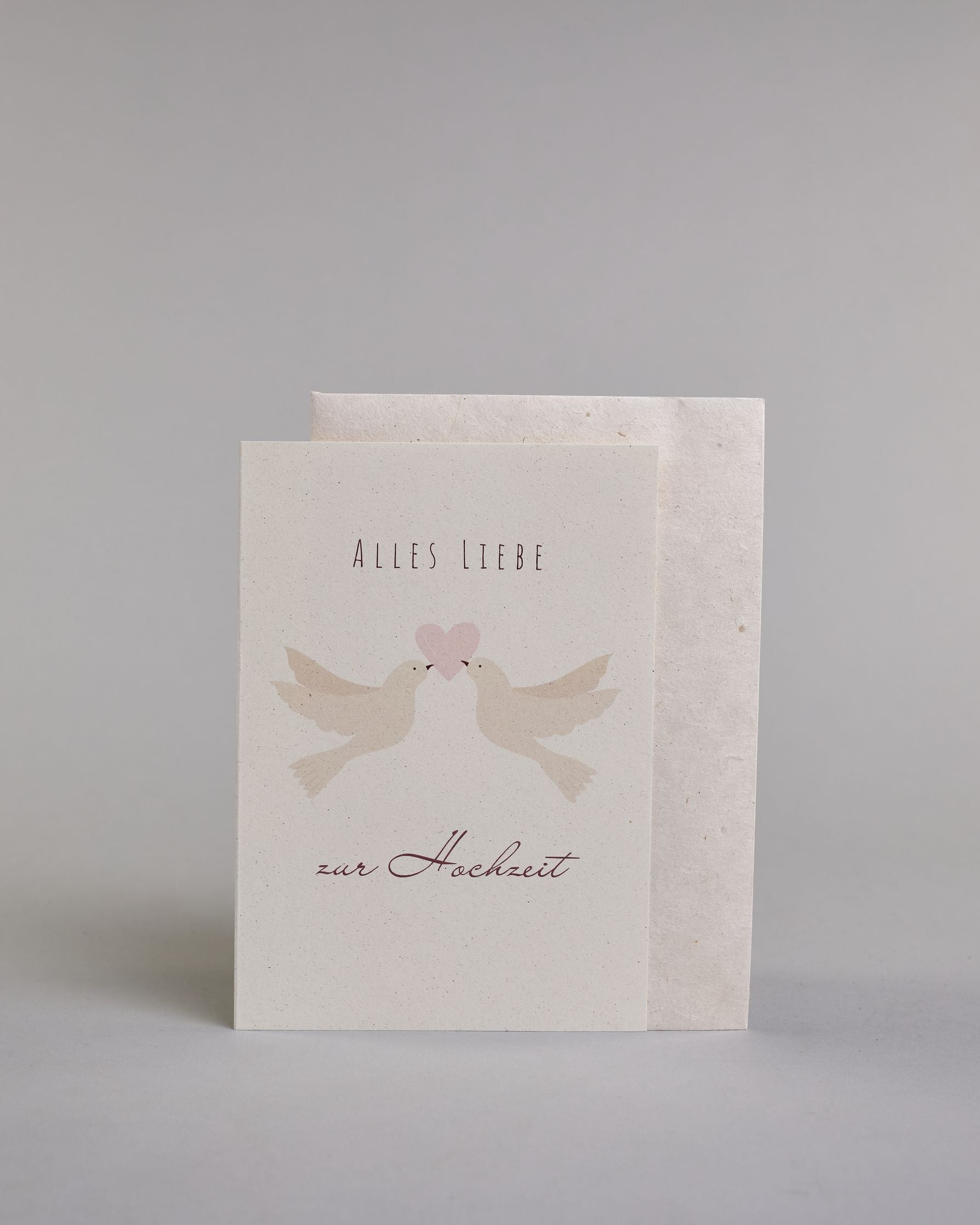 Glückwunschkarte zur Hochzeit mit bordeauxfarbener Kalligrafieaufschrift "Alles Liebe zur Hochzeit". Zwischen den Zeilen "ALLES LIEBE", welche in dünner Schrift gestaltet und der Zeile "zur Hochzeit" welche in geschwungener Schreibschrift gemacht wurde, befinden sich zwei weiße Tauben die gemeinsam mit ihren Schnäbeln ein zartrosanes Herz tragen. Alles ist auf hellem Hintergrund. Dazu ein weißes handgeschöpftes Kuvert. #2