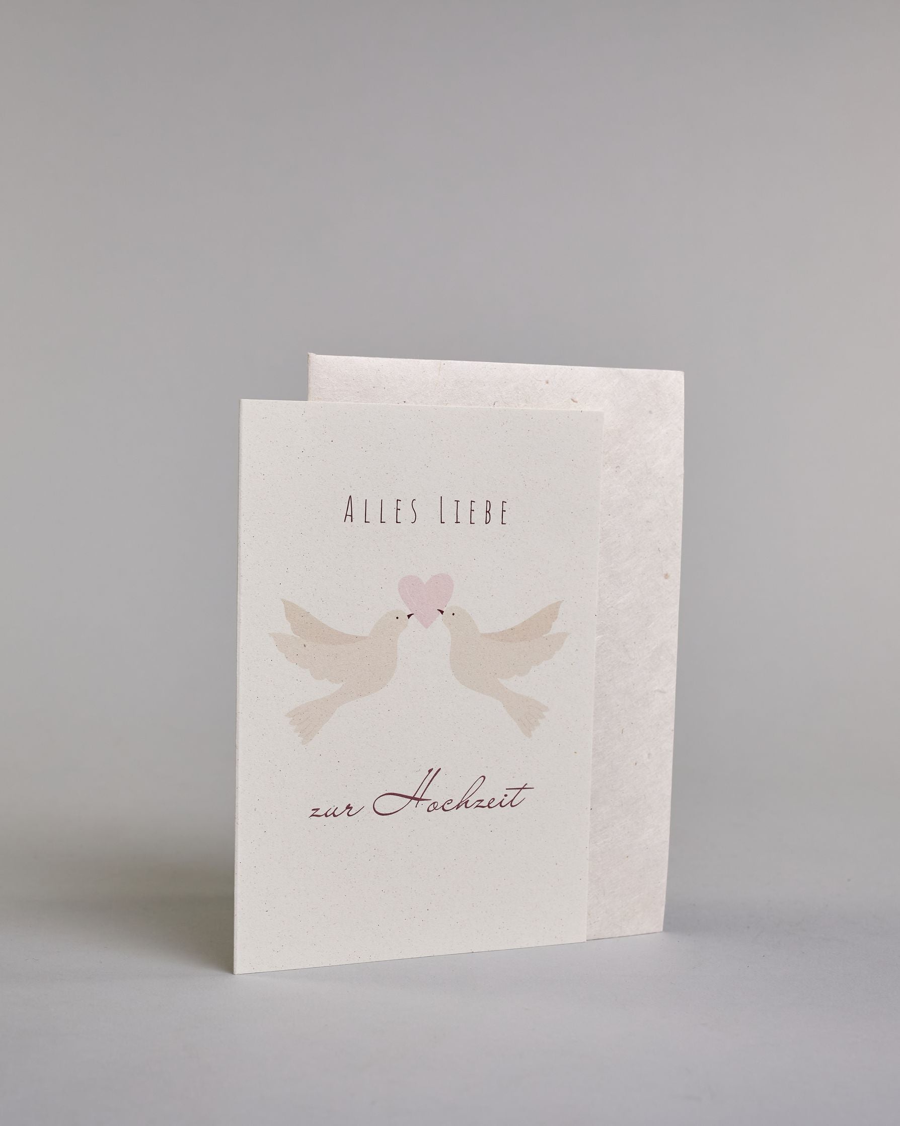 Glückwunschkarte zur Hochzeit mit bordeauxfarbener Kalligrafieaufschrift "Alles Liebe zur Hochzeit". Zwischen den Zeilen "ALLES LIEBE", welche in dünner Schrift gestaltet und der Zeile "zur Hochzeit" welche in geschwungener Schreibschrift gemacht wurde, befinden sich zwei weiße Tauben die gemeinsam mit ihren Schnäbeln ein zartrosanes Herz tragen. Alles ist auf hellem Hintergrund. Dazu ein weißes handgeschöpftes Kuvert. #3