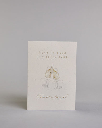 Glückwunschkarte zur Hochzeit mit hellbrauner Kalligrafieaufschrift "Hand in Hand ein Leben lang cheers to forever!". Die Karte ist wiefolgt gegliedert: "HAND IN HAND", darunter "EIN LEBEN LANG", beide Zeilen sind in einer dünnen aufrechten Kalligrafieschrift gestaltet. Darunter stoßen zwei gezeichnete Champagnergläser an, um dessen Gläserhälsen jeweils eine weiße Schleife gebunden ist. Der Champagner schwapt durch den Schwung etwas über den Glasrand und ist mit Golddeteils verfeinert. Darunter ist in gesc…