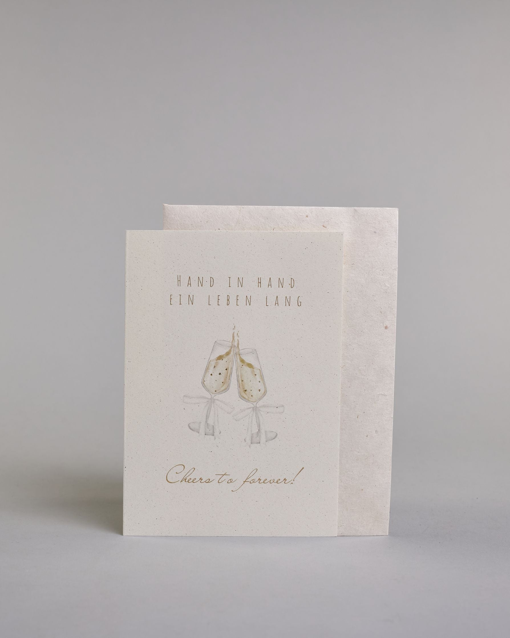 Glückwunschkarte zur Hochzeit mit hellbrauner Kalligrafieaufschrift "Hand in Hand ein Leben lang cheers to forever!". Die Karte ist wiefolgt gegliedert: "HAND IN HAND", darunter "EIN LEBEN LANG", beide Zeilen sind in einer dünnen aufrechten Kalligrafieschrift gestaltet. Darunter stoßen zwei gezeichnete Champagnergläser an, um dessen Gläserhälsen jeweils eine weiße Schleife gebunden ist. Der Champagner schwapt durch den Schwung etwas über den Glasrand und ist mit Golddeteils verfeinert. Darunter ist in gesc…
