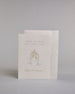 Glückwunschkarte zur Hochzeit mit hellbrauner Kalligrafieaufschrift "Hand in Hand ein Leben lang cheers to forever!". Die Karte ist wiefolgt gegliedert: "HAND IN HAND", darunter "EIN LEBEN LANG", beide Zeilen sind in einer dünnen aufrechten Kalligrafieschrift gestaltet. Darunter stoßen zwei gezeichnete Champagnergläser an, um dessen Gläserhälsen jeweils eine weiße Schleife gebunden ist. Der Champagner schwapt durch den Schwung etwas über den Glasrand und ist mit Golddeteils verfeinert. Darunter ist in gesc…