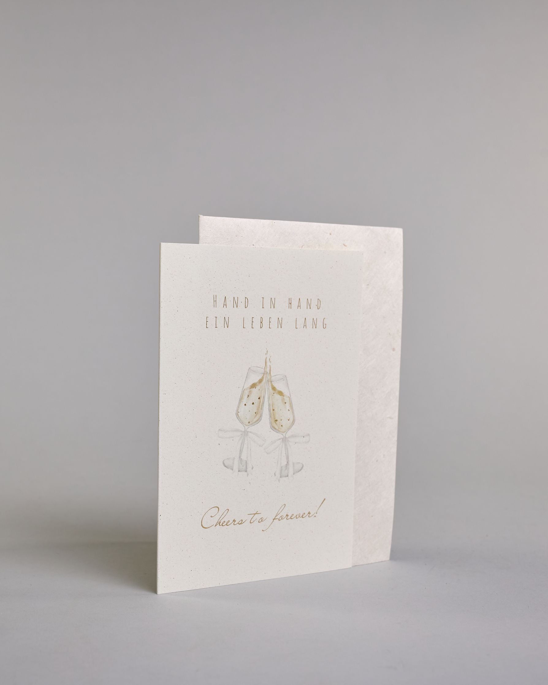 Glückwunschkarte zur Hochzeit mit hellbrauner Kalligrafieaufschrift "Hand in Hand ein Leben lang cheers to forever!". Die Karte ist wiefolgt gegliedert: "HAND IN HAND", darunter "EIN LEBEN LANG", beide Zeilen sind in einer dünnen aufrechten Kalligrafieschrift gestaltet. Darunter stoßen zwei gezeichnete Champagnergläser an, um dessen Gläserhälsen jeweils eine weiße Schleife gebunden ist. Der Champagner schwapt durch den Schwung etwas über den Glasrand und ist mit Golddeteils verfeinert. Darunter ist in gesc…
