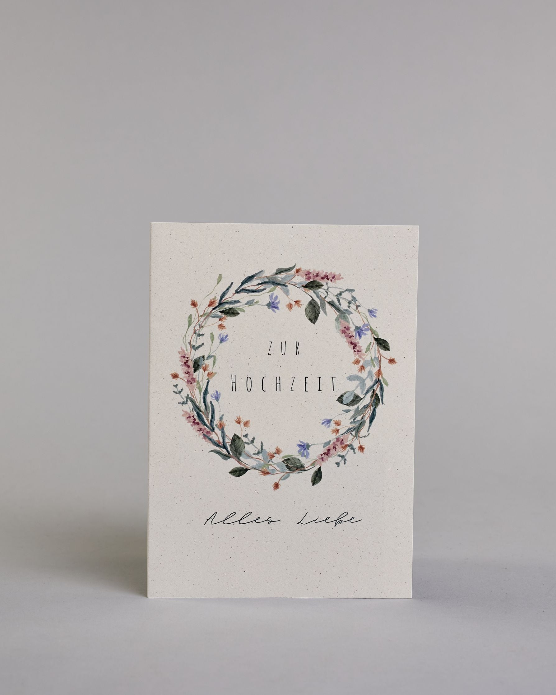 Glückwunschkarte zur Hochzeit mit schwarzer Kalligrafieaufschrift "Zur Hochzeit Alles Liebe". Die Worte "ZUR HOCHZEIT" sind auf zwei Zeilen aufgeteilt und befinden sich in einem Kranz aus rosanen und blauen Wildblumen und grünen und roten Zweigen und Blättern.  Die Schrift ist eine dünne aufrechte Kalligrafieschrift. Unter dem Kranz befinden sich die Worte "Alles Liebe" in schwarzer geschwungener Schreibschritf. Alles ist auf hellem Hintergrund. Dazu ein weißes handgeschöpftes Kuvert. #1