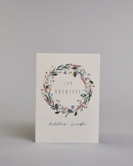 Glückwunschkarte zur Hochzeit mit schwarzer Kalligrafieaufschrift "Zur Hochzeit Alles Liebe". Die Worte "ZUR HOCHZEIT" sind auf zwei Zeilen aufgeteilt und befinden sich in einem Kranz aus rosanen und blauen Wildblumen und grünen und roten Zweigen und Blättern.  Die Schrift ist eine dünne aufrechte Kalligrafieschrift. Unter dem Kranz befinden sich die Worte "Alles Liebe" in schwarzer geschwungener Schreibschritf. Alles ist auf hellem Hintergrund. Dazu ein weißes handgeschöpftes Kuvert. #1