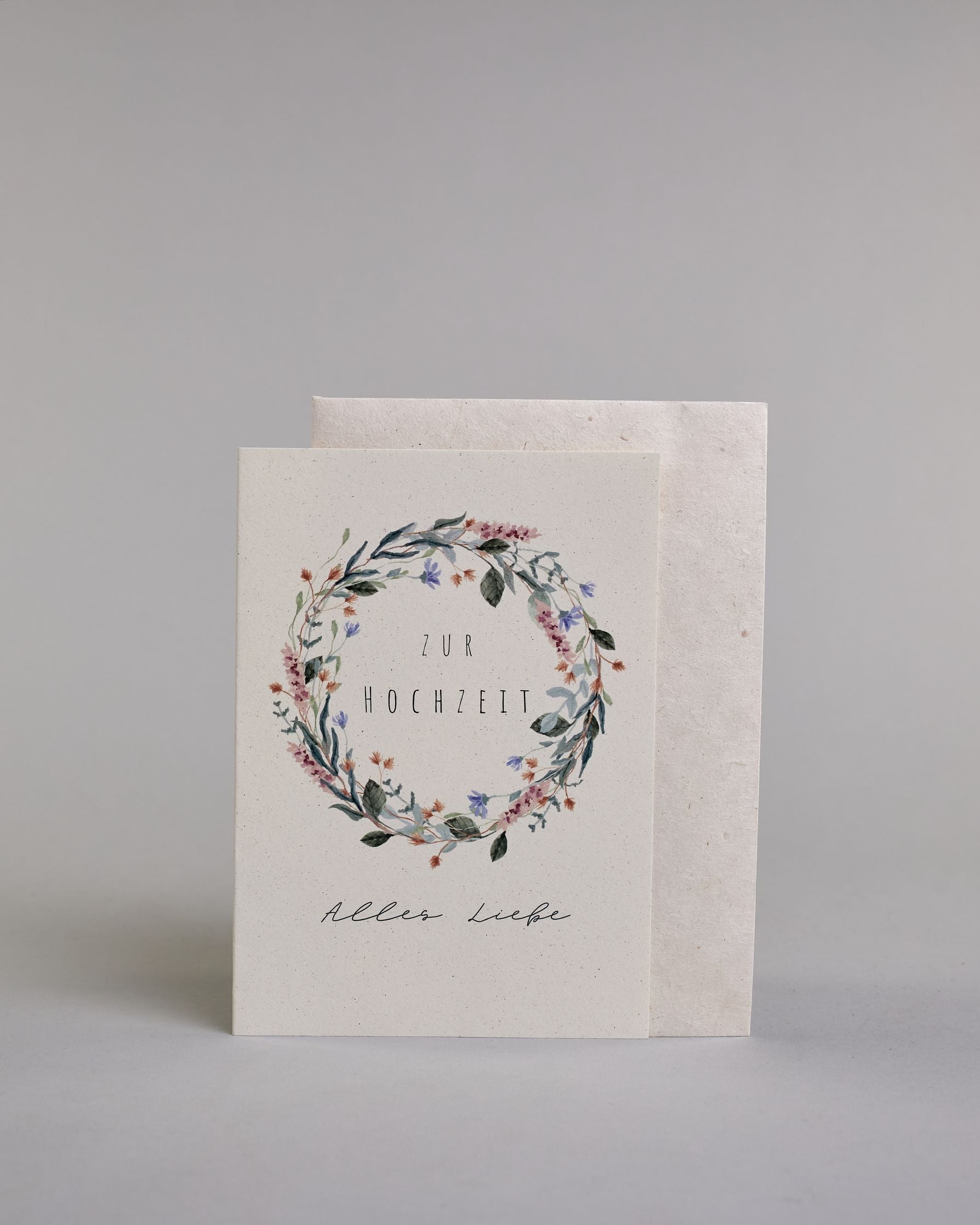 Glückwunschkarte zur Hochzeit mit schwarzer Kalligrafieaufschrift "Zur Hochzeit Alles Liebe". Die Worte "ZUR HOCHZEIT" sind auf zwei Zeilen aufgeteilt und befinden sich in einem Kranz aus rosanen und blauen Wildblumen und grünen und roten Zweigen und Blättern.  Die Schrift ist eine dünne aufrechte Kalligrafieschrift. Unter dem Kranz befinden sich die Worte "Alles Liebe" in schwarzer geschwungener Schreibschritf. Alles ist auf hellem Hintergrund. Dazu ein weißes handgeschöpftes Kuvert. #2