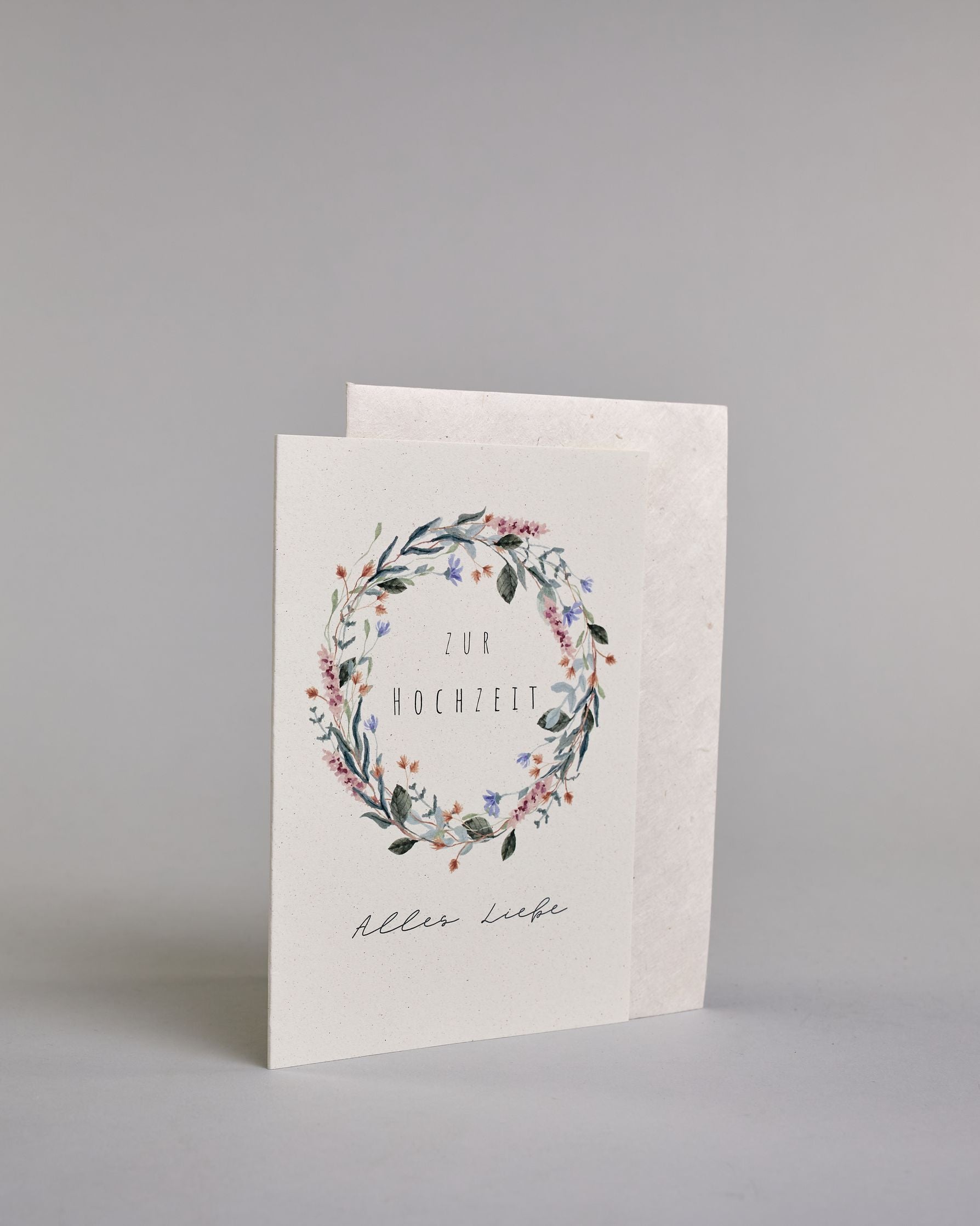 Glückwunschkarte zur Hochzeit mit schwarzer Kalligrafieaufschrift "Zur Hochzeit Alles Liebe". Die Worte "ZUR HOCHZEIT" sind auf zwei Zeilen aufgeteilt und befinden sich in einem Kranz aus rosanen und blauen Wildblumen und grünen und roten Zweigen und Blättern.  Die Schrift ist eine dünne aufrechte Kalligrafieschrift. Unter dem Kranz befinden sich die Worte "Alles Liebe" in schwarzer geschwungener Schreibschritf. Alles ist auf hellem Hintergrund. Dazu ein weißes handgeschöpftes Kuvert. #3