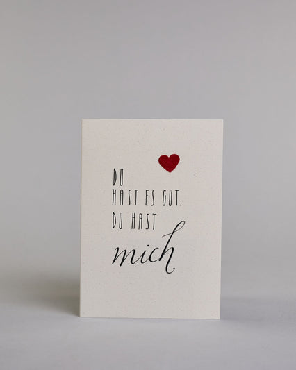 Klappkarte für Liebende mit schwarzer Kalligrafieaufschrift "Du hast es gut. Du hast mich" mit einem rotem aufgeklebtem Herz auf hellem Hintergrund. Die Schrift ist auf 4 Zeilen aufgeteilt. "DU", darunter "HAST ES GUT.", darunter "DU HAST" alles in einer dünnen geraden Kalligrafieschrift, darunter "mich" in einer geschwungenen Schreibschrift. Die Schrift ist eher linksbündig, das Herz klebt auf Höhe der ersten Zeile, mit dem "DU" rechts auf der Karte.  Dazu ein rot-weißes handgeschöpftes Kuvert. #1