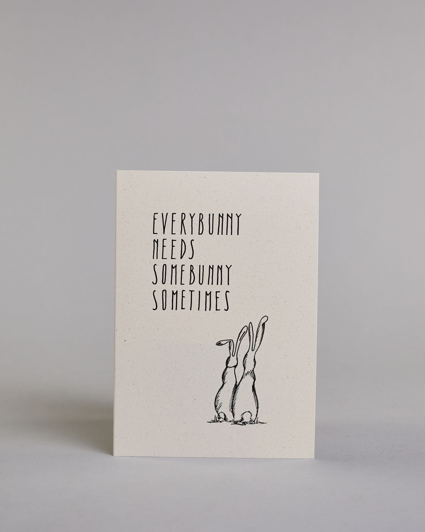 Klappkarte für Liebende auf Englisch mit schwarzer Kalligrafieaufschrift "Everybunny needs somebunny sometimes", darunter sind zwei gezeichnete Hasen von hinten auf hellem Hintergrund zu sehen. Die Kalligrafieschrift ist dünn und gerade und teilt sich in vier Zeilen mit jeweils immer einem Wort auf. Die Schrift ist im oberen linken Teil der Karte, während die Hasen rechts unten sind. Dazu ein grün-goldenes handgeschöpftes Kuvert. #1