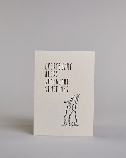 Klappkarte für Liebende auf Englisch mit schwarzer Kalligrafieaufschrift "Everybunny needs somebunny sometimes", darunter sind zwei gezeichnete Hasen von hinten auf hellem Hintergrund zu sehen. Die Kalligrafieschrift ist dünn und gerade und teilt sich in vier Zeilen mit jeweils immer einem Wort auf. Die Schrift ist im oberen linken Teil der Karte, während die Hasen rechts unten sind. Dazu ein grün-goldenes handgeschöpftes Kuvert. #1