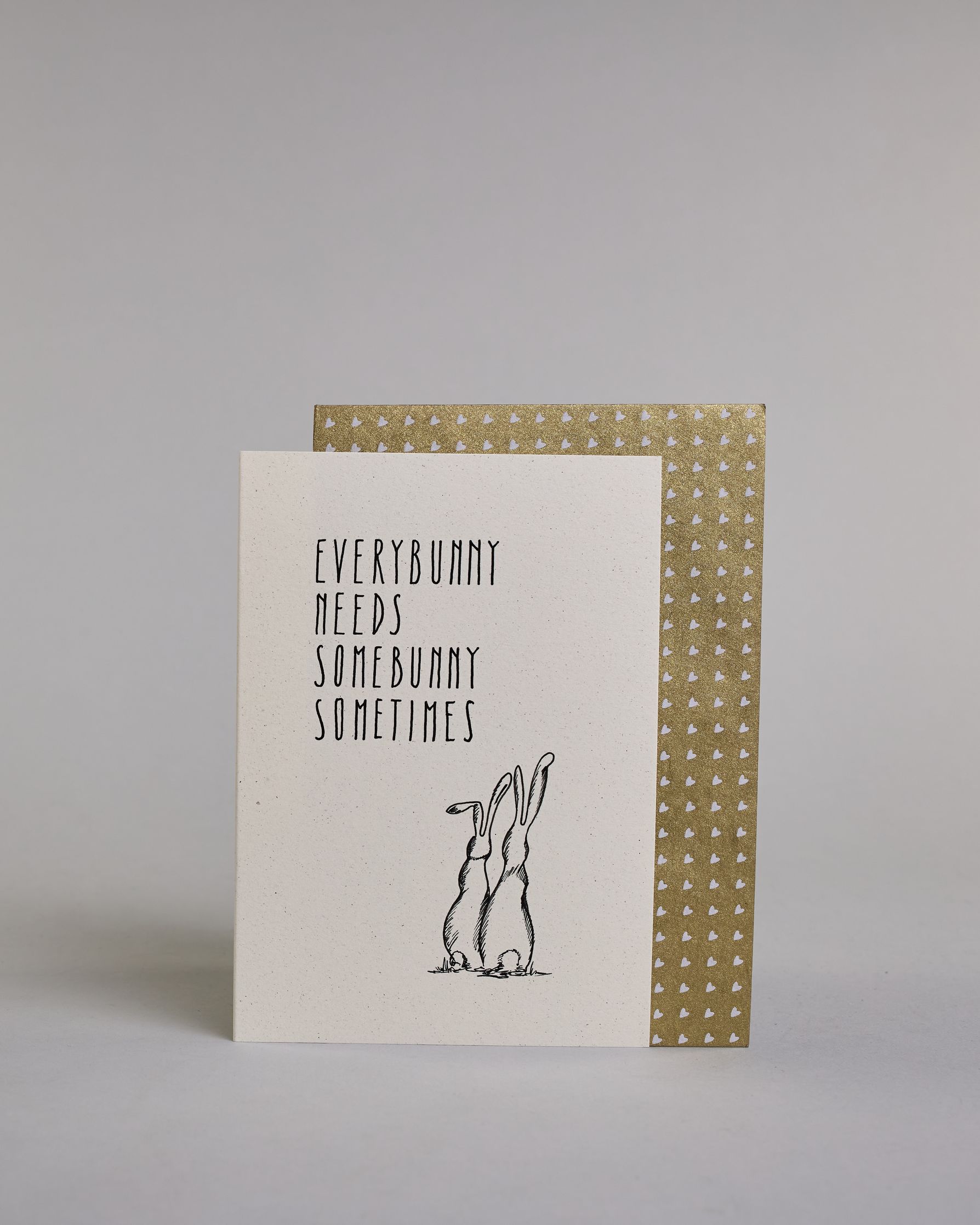 Klappkarte für Liebende auf Englisch mit schwarzer Kalligrafieaufschrift "Everybunny needs somebunny sometimes", darunter sind zwei gezeichnete Hasen von hinten auf hellem Hintergrund zu sehen. Die Kalligrafieschrift ist dünn und gerade und teilt sich in vier Zeilen mit jeweils immer einem Wort auf. Die Schrift ist im oberen linken Teil der Karte, während die Hasen rechts unten sind. Dazu ein grün-goldenes handgeschöpftes Kuvert. #2
