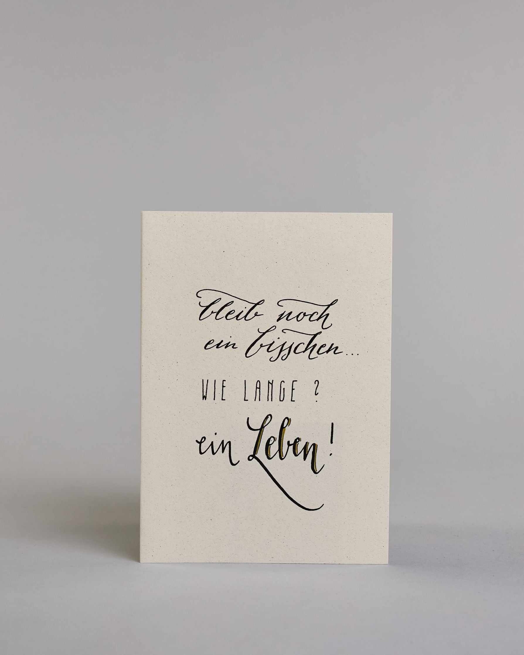 Klappkarte für Liebende mit schwarzer Kalligrafieaufschrift "Bleib noch ein bisschen... Wie lange? Ein Leben!" auf hellem Hintergrund. Der Text ist auf vier Zeilen mit jeweils immer zwei Worten auf geteilt. Die Zeilen "bleib noch", "ein bisschen" und "ein Leben!" sind in dünner geschwungener Schreibschrift gestaltet, während "WIE LANGE?" in dünner, gerader Kalligrafieschrift geschrieben ist. Dazu ein grün-goldenes handgeschöpftes Kuvert. #1