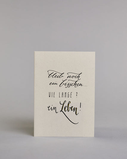 Klappkarte für Liebende mit schwarzer Kalligrafieaufschrift "Bleib noch ein bisschen... Wie lange? Ein Leben!" auf hellem Hintergrund. Der Text ist auf vier Zeilen mit jeweils immer zwei Worten auf geteilt. Die Zeilen "bleib noch", "ein bisschen" und "ein Leben!" sind in dünner geschwungener Schreibschrift gestaltet, während "WIE LANGE?" in dünner, gerader Kalligrafieschrift geschrieben ist. Dazu ein grün-goldenes handgeschöpftes Kuvert. #1
