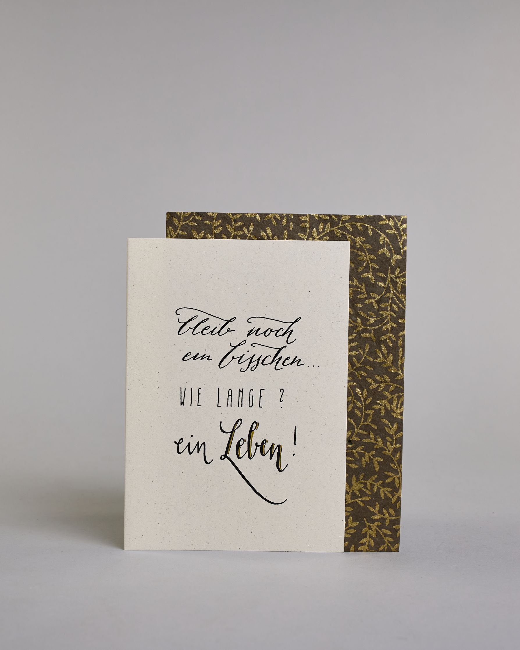 Klappkarte für Liebende mit schwarzer Kalligrafieaufschrift "Bleib noch ein bisschen... Wie lange? Ein Leben!" auf hellem Hintergrund. Der Text ist auf vier Zeilen mit jeweils immer zwei Worten auf geteilt. Die Zeilen "bleib noch", "ein bisschen" und "ein Leben!" sind in dünner geschwungener Schreibschrift gestaltet, während "WIE LANGE?" in dünner, gerader Kalligrafieschrift geschrieben ist. Dazu ein grün-goldenes handgeschöpftes Kuvert. #2