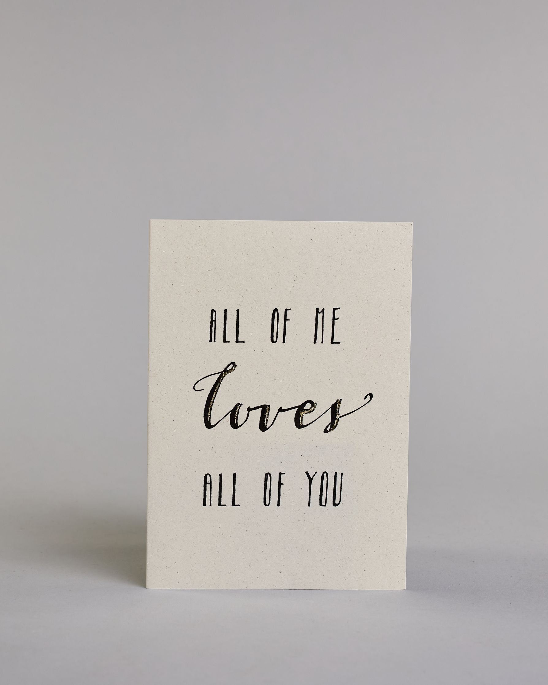 Klappkarte für Liebende auf Englisch mit schwarzer Kalligrafieaufschrift "All of me love all of you" auf hellem Hintergrund. Der Text ist in folgende drei Zeilen aufgeteilt: "ALL OF ME" in dünner gerader Kalligrafieschrift, darunter "loves" in geschwungener Schreibschrift und goldglitzernden Verzierungen, darunter "ALL OF YOU" wieder in gerader, dünner Schrift. Dazu ein weiß-goldenes handgeschöpftes Kuvert. #1