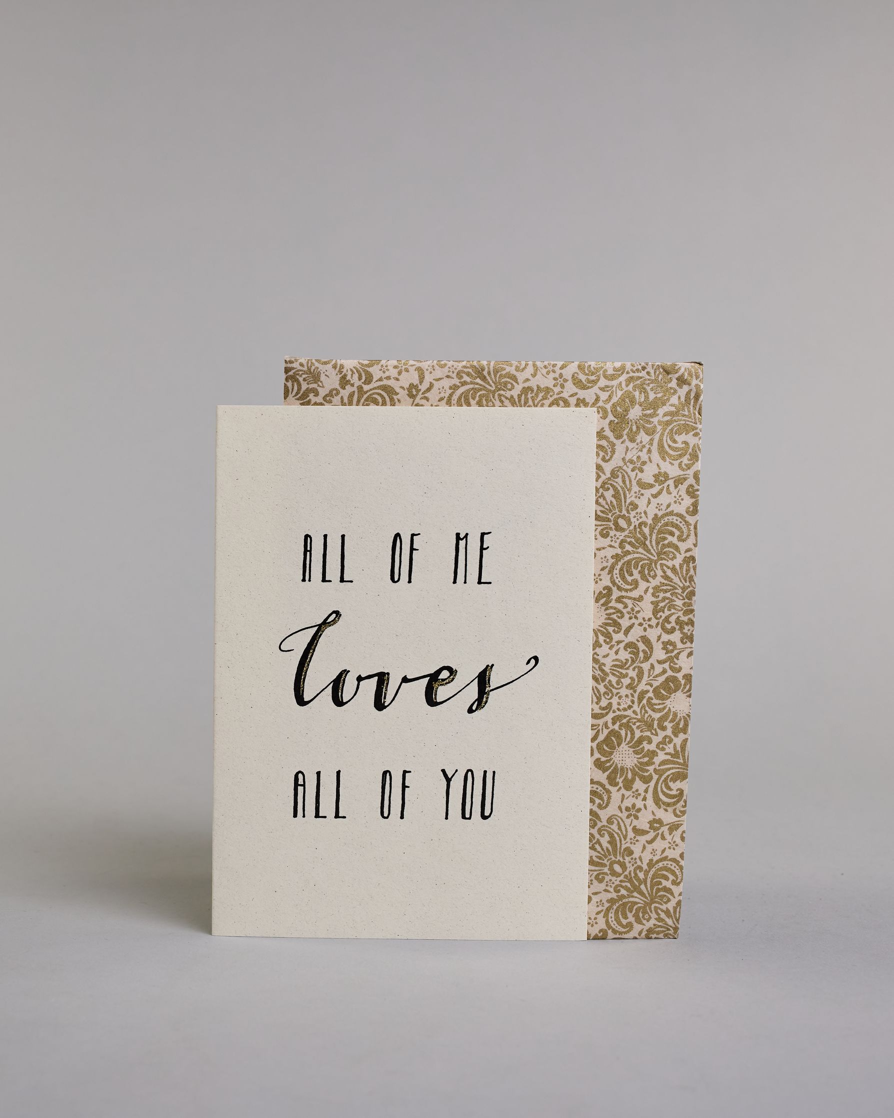 Klappkarte für Liebende auf Englisch mit schwarzer Kalligrafieaufschrift "All of me love all of you" auf hellem Hintergrund. Der Text ist in folgende drei Zeilen aufgeteilt: "ALL OF ME" in dünner gerader Kalligrafieschrift, darunter "loves" in geschwungener Schreibschrift und goldglitzernden Verzierungen, darunter "ALL OF YOU" wieder in gerader, dünner Schrift. Dazu ein weiß-goldenes handgeschöpftes Kuvert. #2