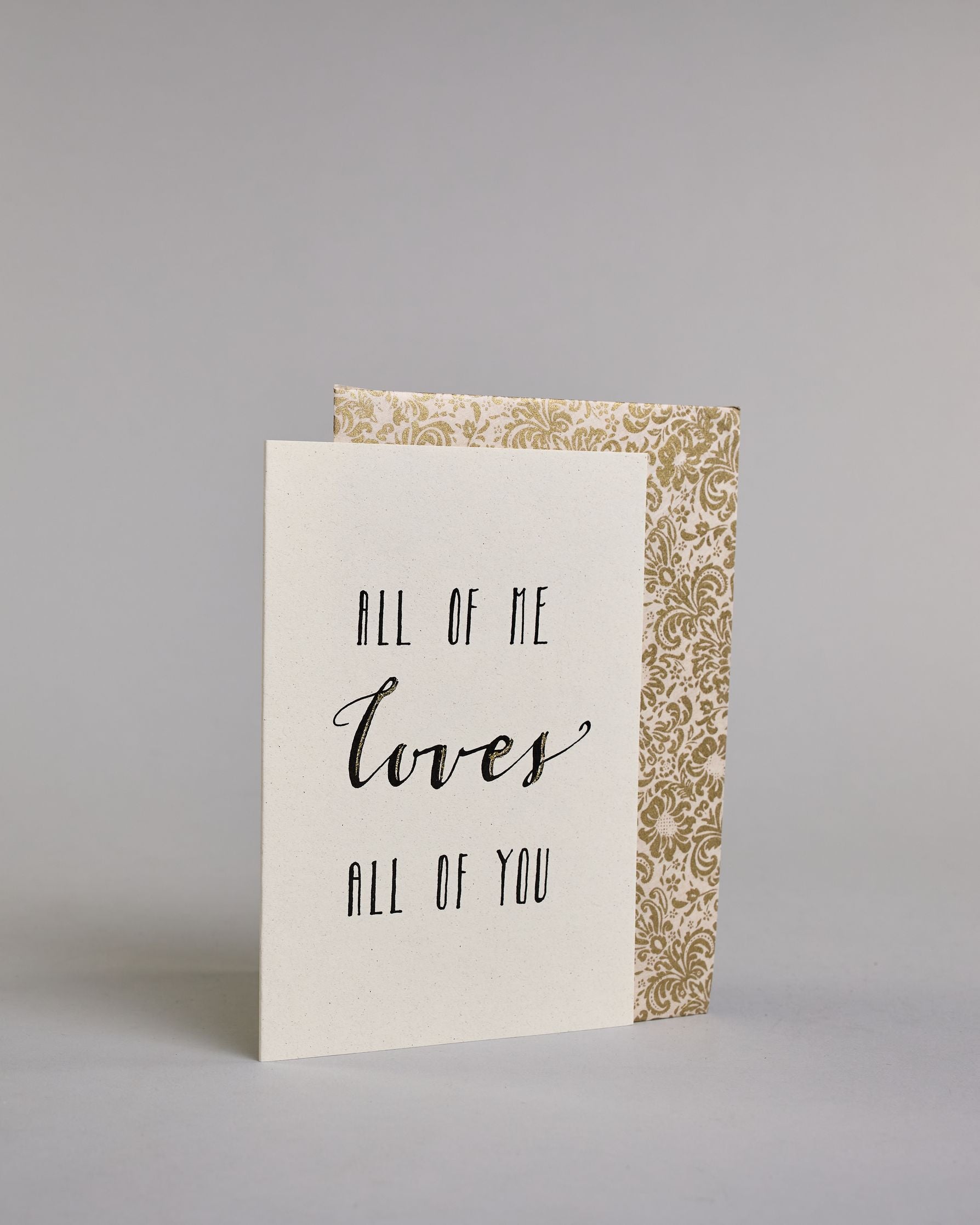 Klappkarte für Liebende auf Englisch mit schwarzer Kalligrafieaufschrift "All of me love all of you" auf hellem Hintergrund. Der Text ist in folgende drei Zeilen aufgeteilt: "ALL OF ME" in dünner gerader Kalligrafieschrift, darunter "loves" in geschwungener Schreibschrift und goldglitzernden Verzierungen, darunter "ALL OF YOU" wieder in gerader, dünner Schrift. Dazu ein weiß-goldenes handgeschöpftes Kuvert. #3