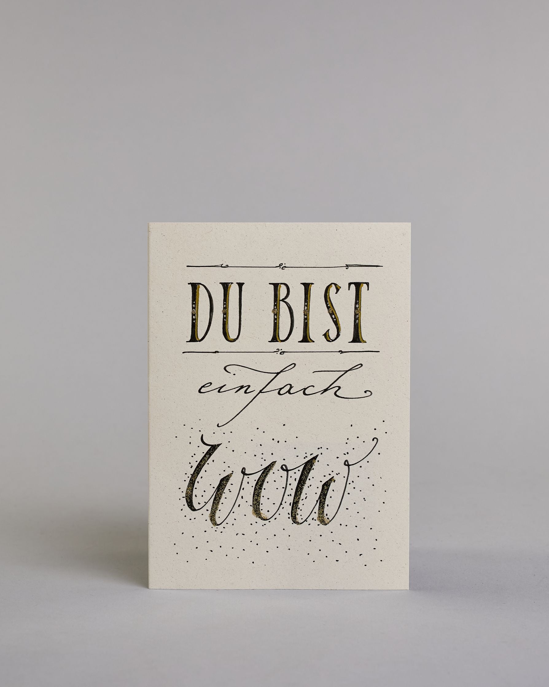 Klappkarte für Liebende mit schwarzer Kalligrafieaufschrift "Du bist einfach wow" auf hellem Hintergrund. Der Text ist in folgende drei Zeilen aufgeteilt: "DU BIST" in breiter gerader Kalligrafieschrift darüber und darunter eine dekorative horizontale Linie, darunter "einfach" in geschwungener Schreibschrift, darunter "wow" in einer anderen geschwungenen Schreibschrift dahinter Pünktchen. Dazu ein handgeschöpftes Kuvert. #1