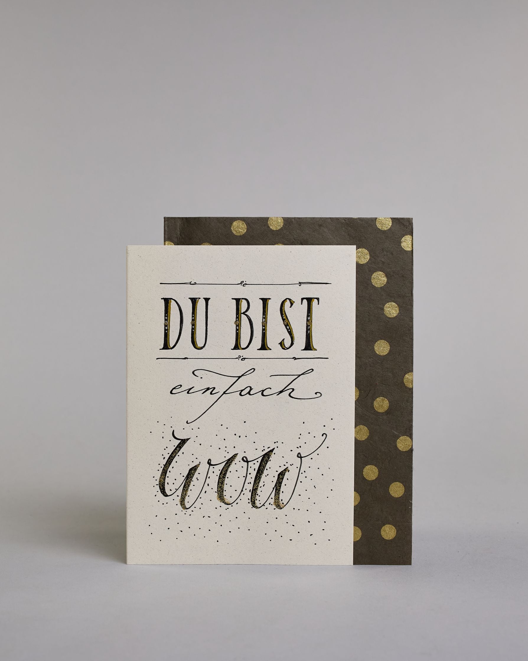 Klappkarte für Liebende mit schwarzer Kalligrafieaufschrift "Du bist einfach wow" auf hellem Hintergrund. Der Text ist in folgende drei Zeilen aufgeteilt: "DU BIST" in breiter gerader Kalligrafieschrift darüber und darunter eine dekorative horizontale Linie, darunter "einfach" in geschwungener Schreibschrift, darunter "wow" in einer anderen geschwungenen Schreibschrift dahinter Pünktchen. Dazu ein handgeschöpftes Kuvert. #2