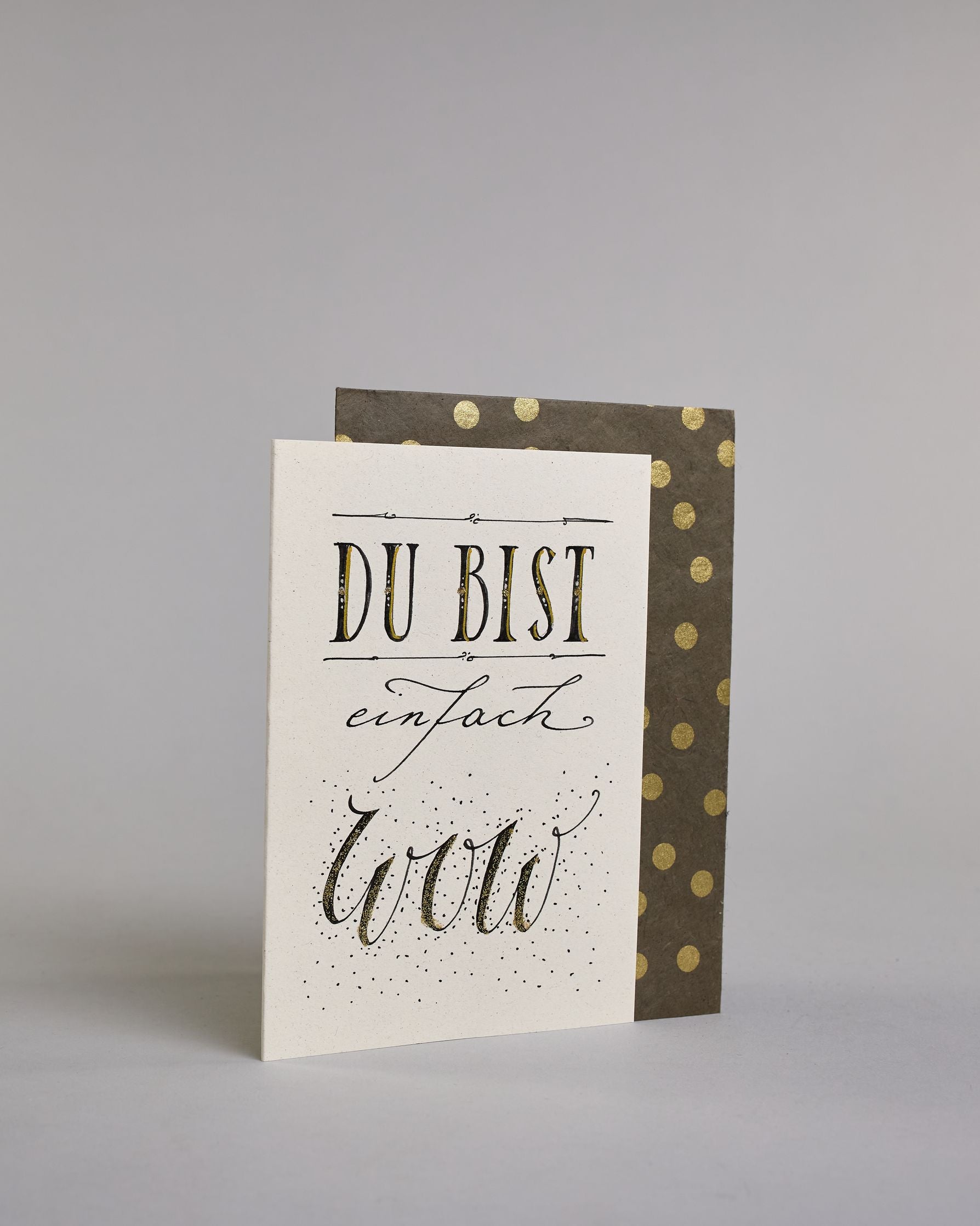 Klappkarte für Liebende mit schwarzer Kalligrafieaufschrift "Du bist einfach wow" auf hellem Hintergrund. Der Text ist in folgende drei Zeilen aufgeteilt: "DU BIST" in breiter gerader Kalligrafieschrift darüber und darunter eine dekorative horizontale Linie, darunter "einfach" in geschwungener Schreibschrift, darunter "wow" in einer anderen geschwungenen Schreibschrift dahinter Pünktchen. Dazu ein handgeschöpftes Kuvert. #3