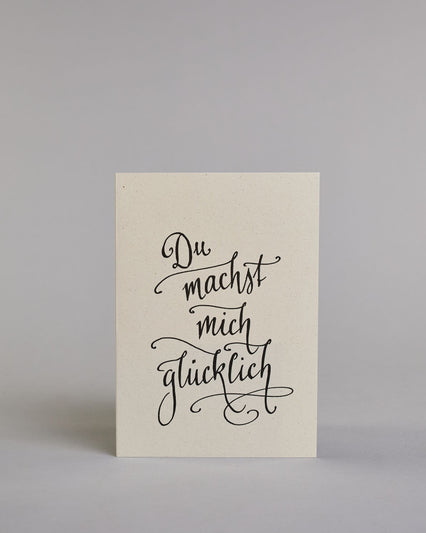 Klappkarte für Liebende mit schwarzer Kalligrafieaufschrift "Du machst mich glücklich" auf hellem Hintergrund. Der Text ist in vier Zeilen mit jeweils einem Wort aufgeteilt und in einer sehr schwungvollen dünnen Kalligrafieschrift gestaltet. Dazu ein gold-weißes handgeschöpftes Kuvert. #1