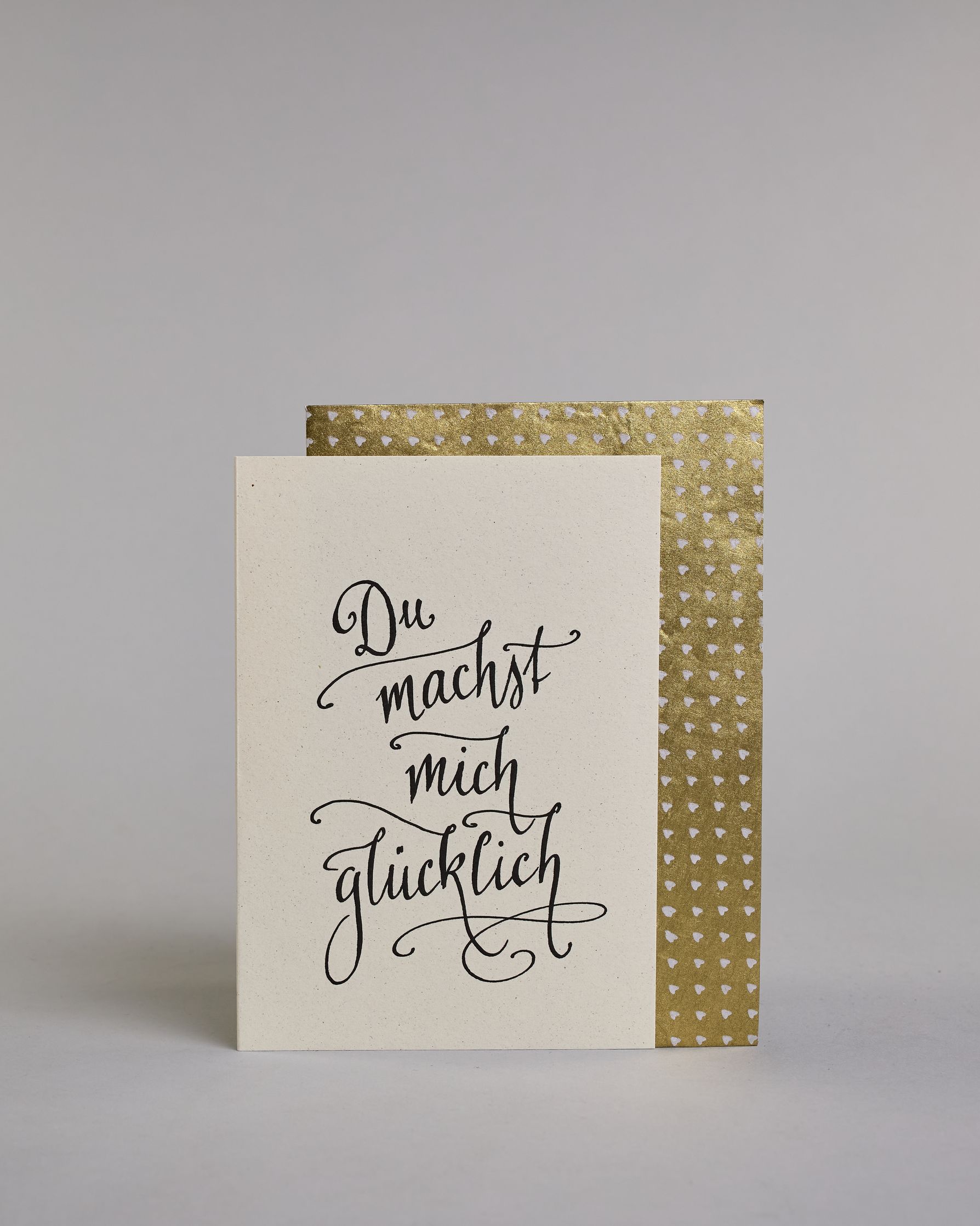 Klappkarte für Liebende mit schwarzer Kalligrafieaufschrift "Du machst mich glücklich" auf hellem Hintergrund. Der Text ist in vier Zeilen mit jeweils einem Wort aufgeteilt und in einer sehr schwungvollen dünnen Kalligrafieschrift gestaltet. Dazu ein gold-weißes handgeschöpftes Kuvert. #2