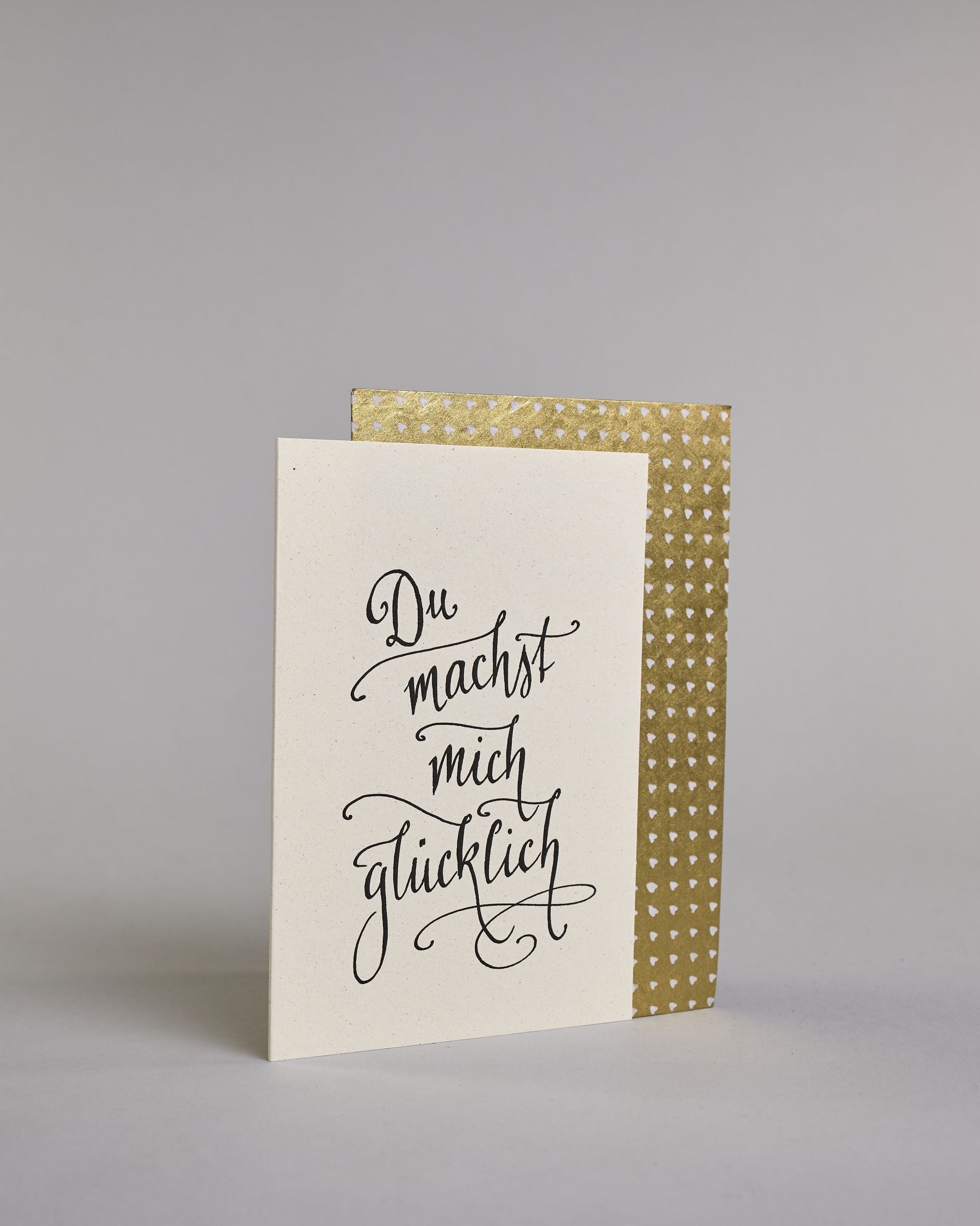 Klappkarte für Liebende mit schwarzer Kalligrafieaufschrift "Du machst mich glücklich" auf hellem Hintergrund. Der Text ist in vier Zeilen mit jeweils einem Wort aufgeteilt und in einer sehr schwungvollen dünnen Kalligrafieschrift gestaltet. Dazu ein gold-weißes handgeschöpftes Kuvert. #3
