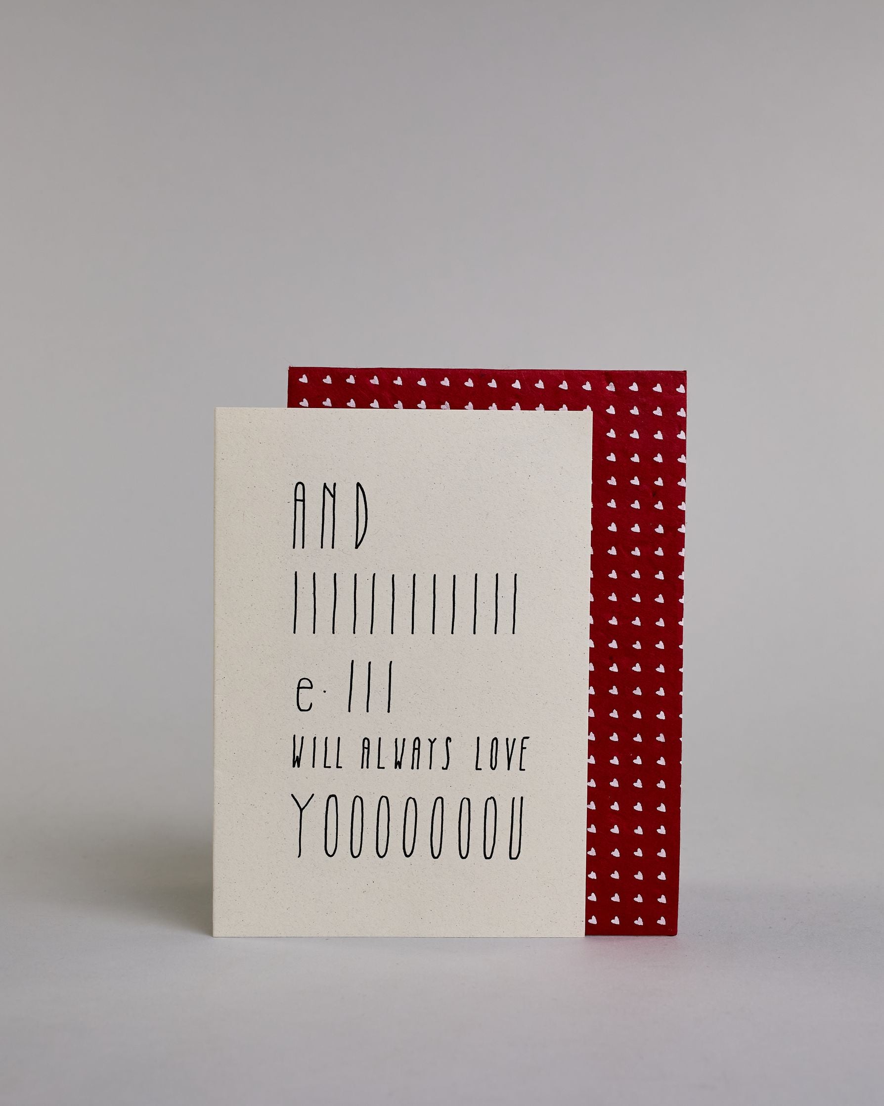 Klappkarte für Liebende auf Englisch mit schwarzer Kalligrafieaufschrift "And I will always love you" auf hellem Hintergrund. Der Text ist in folgende fünf Zeilen aufgeteilt: "AND", darunter "IIIIIIIIIIII", darunter "e-III", darunter "WILL ALWAYS LOVE", darunter "YOOOOOOOU" alle Zeilen sind in der gleichen dünnen geraden Kalligrafieschrift gestaltet und es ist eine Referenz zu dem Bekannten Whitney Houston Song, daher ist das "I" und das "you" in die Länge gezogen. Dazu ein rot-weißes handgeschöpftes Kuver…