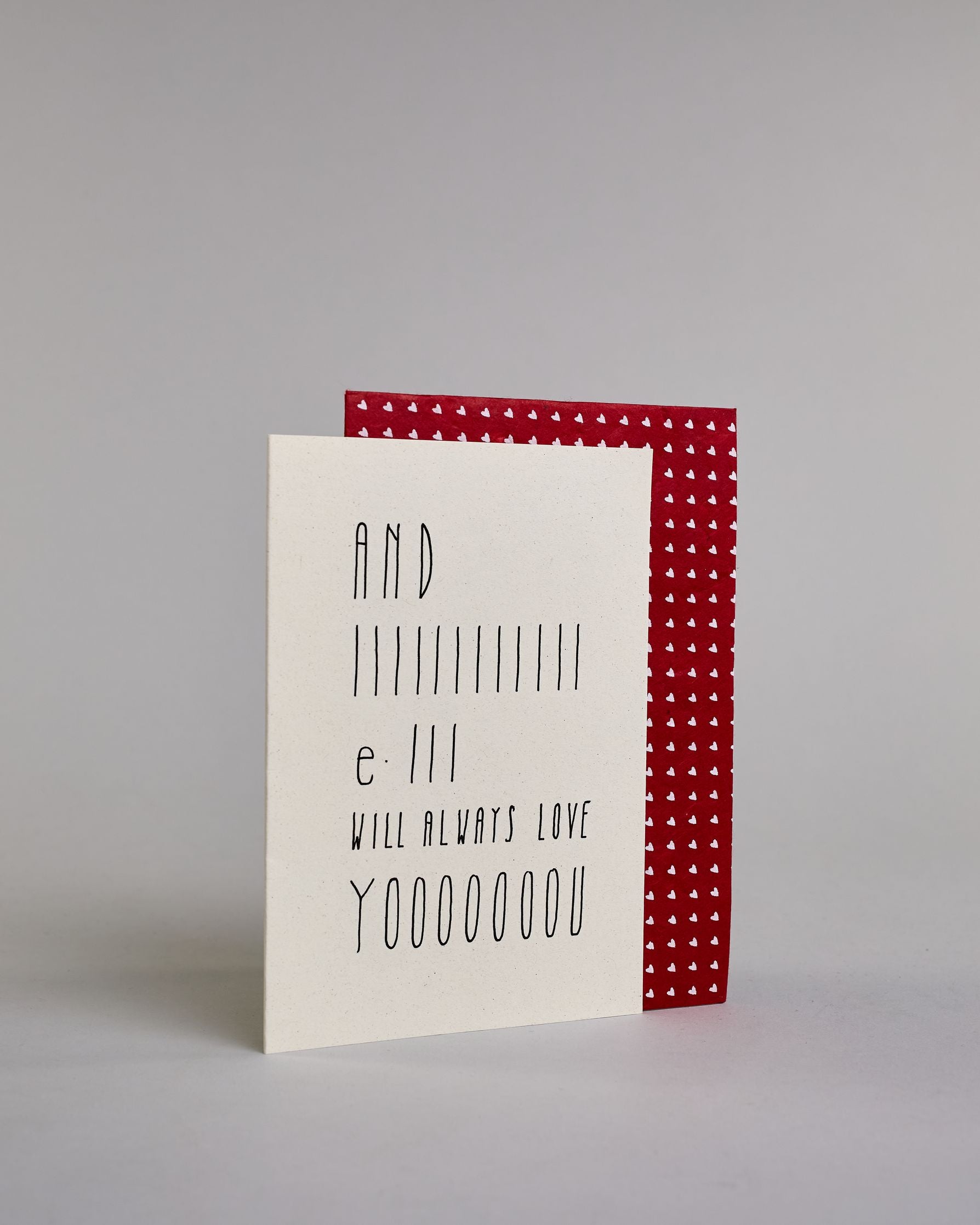 Klappkarte für Liebende auf Englisch mit schwarzer Kalligrafieaufschrift "And I will always love you" auf hellem Hintergrund. Der Text ist in folgende fünf Zeilen aufgeteilt: "AND", darunter "IIIIIIIIIIII", darunter "e-III", darunter "WILL ALWAYS LOVE", darunter "YOOOOOOOU" alle Zeilen sind in der gleichen dünnen geraden Kalligrafieschrift gestaltet und es ist eine Referenz zu dem Bekannten Whitney Houston Song, daher ist das "I" und das "you" in die Länge gezogen. Dazu ein rot-weißes handgeschöpftes Kuver…
