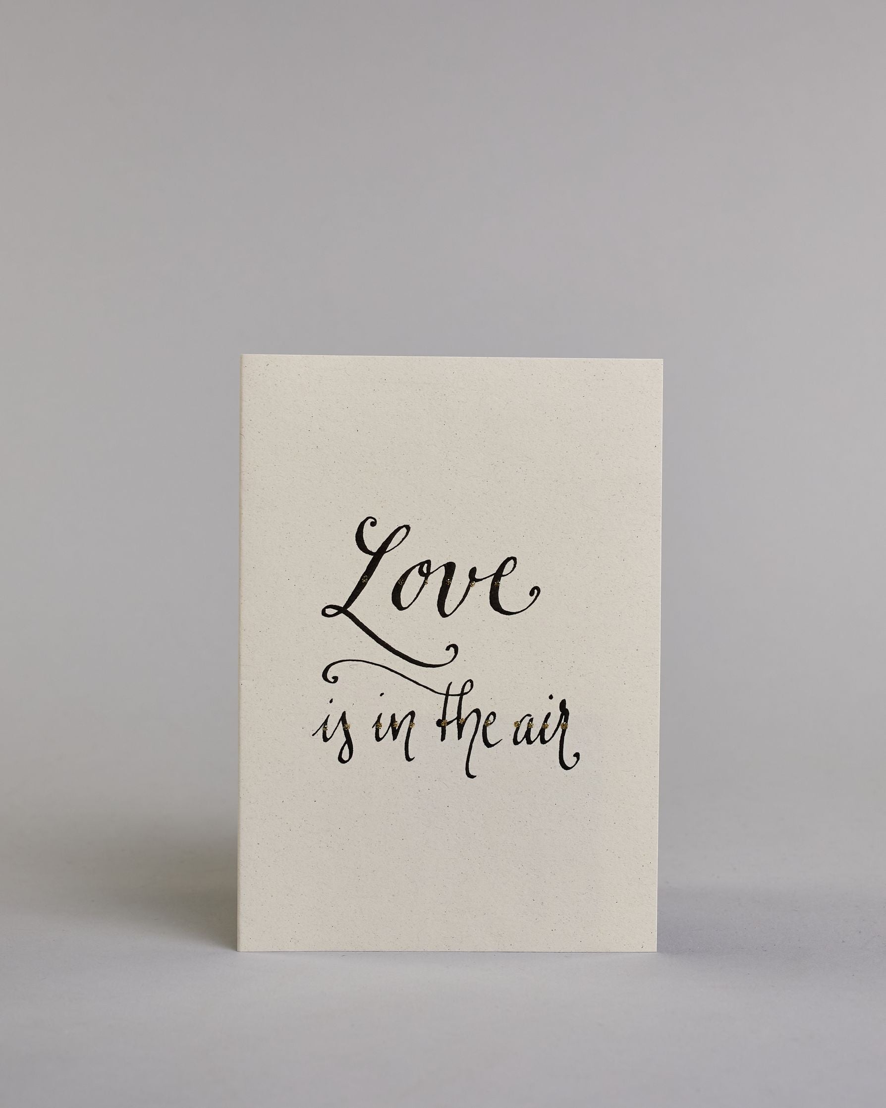 Klappkarte für Liebende auf Englisch mit schwarzer Kalligrafieaufschrift "Love is in the air" auf hellem Hintergrund. Der Text ist in zwei Zeilen aufgeteilt. "Love" und darunter "is in the air". Die Zeilen sind in der gleichen dünnen geschwungenen Schreibschrift gestaltet, wobei das "Love" größer ist als das "is in the air" und es ist eine Referenz zu dem Bekannten John Paul Young Song. Dazu ein rot-weißes handgeschöpftes Kuvert. #1