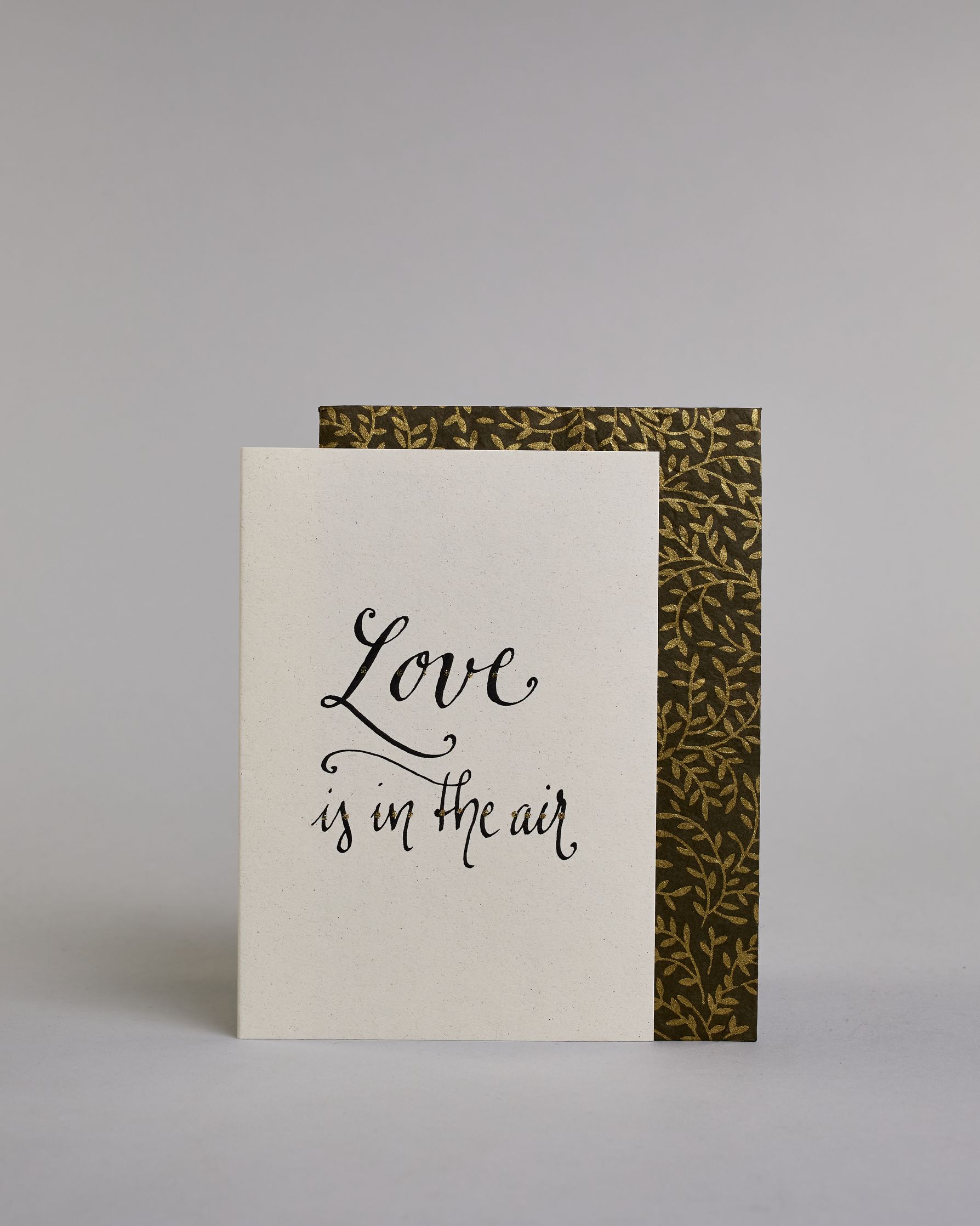 Klappkarte für Liebende auf Englisch mit schwarzer Kalligrafieaufschrift "Love is in the air" auf hellem Hintergrund. Der Text ist in zwei Zeilen aufgeteilt. "Love" und darunter "is in the air". Die Zeilen sind in der gleichen dünnen geschwungenen Schreibschrift gestaltet, wobei das "Love" größer ist als das "is in the air" und es ist eine Referenz zu dem Bekannten John Paul Young Song. Dazu ein rot-weißes handgeschöpftes Kuvert. #2