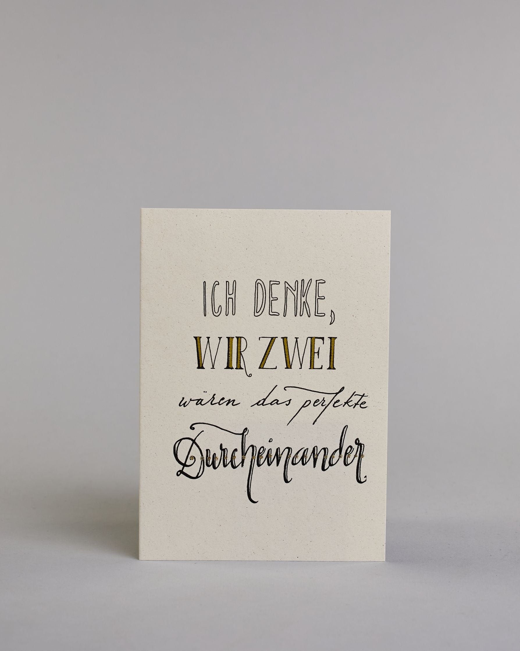 Klappkarte für Liebende mit schwarzer Kalligrafieaufschrift "Ich denke wir zwei wären das perfekte Durcheinander" auf hellem Hintergrund. Der Text ist in folgende vier Zeilen aufgeteilt: "ICH DENKE," in dicken geraden Kalligrafieschrift die weiß ausgemalt ist, darunter "WIR ZWEI" in einer geraden Kalligrafieschrift, darunter "wären das perfekte" in einer dünnen kleinen Schreibschrift, darunter "Durcheinander" in einer größeren Schreibschrift gestaltet . Dazu ein weiß-goldenes handgeschöpftes Kuvert. #1