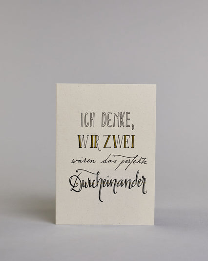 Klappkarte für Liebende mit schwarzer Kalligrafieaufschrift "Ich denke wir zwei wären das perfekte Durcheinander" auf hellem Hintergrund. Der Text ist in folgende vier Zeilen aufgeteilt: "ICH DENKE," in dicken geraden Kalligrafieschrift die weiß ausgemalt ist, darunter "WIR ZWEI" in einer geraden Kalligrafieschrift, darunter "wären das perfekte" in einer dünnen kleinen Schreibschrift, darunter "Durcheinander" in einer größeren Schreibschrift gestaltet . Dazu ein weiß-goldenes handgeschöpftes Kuvert. #1