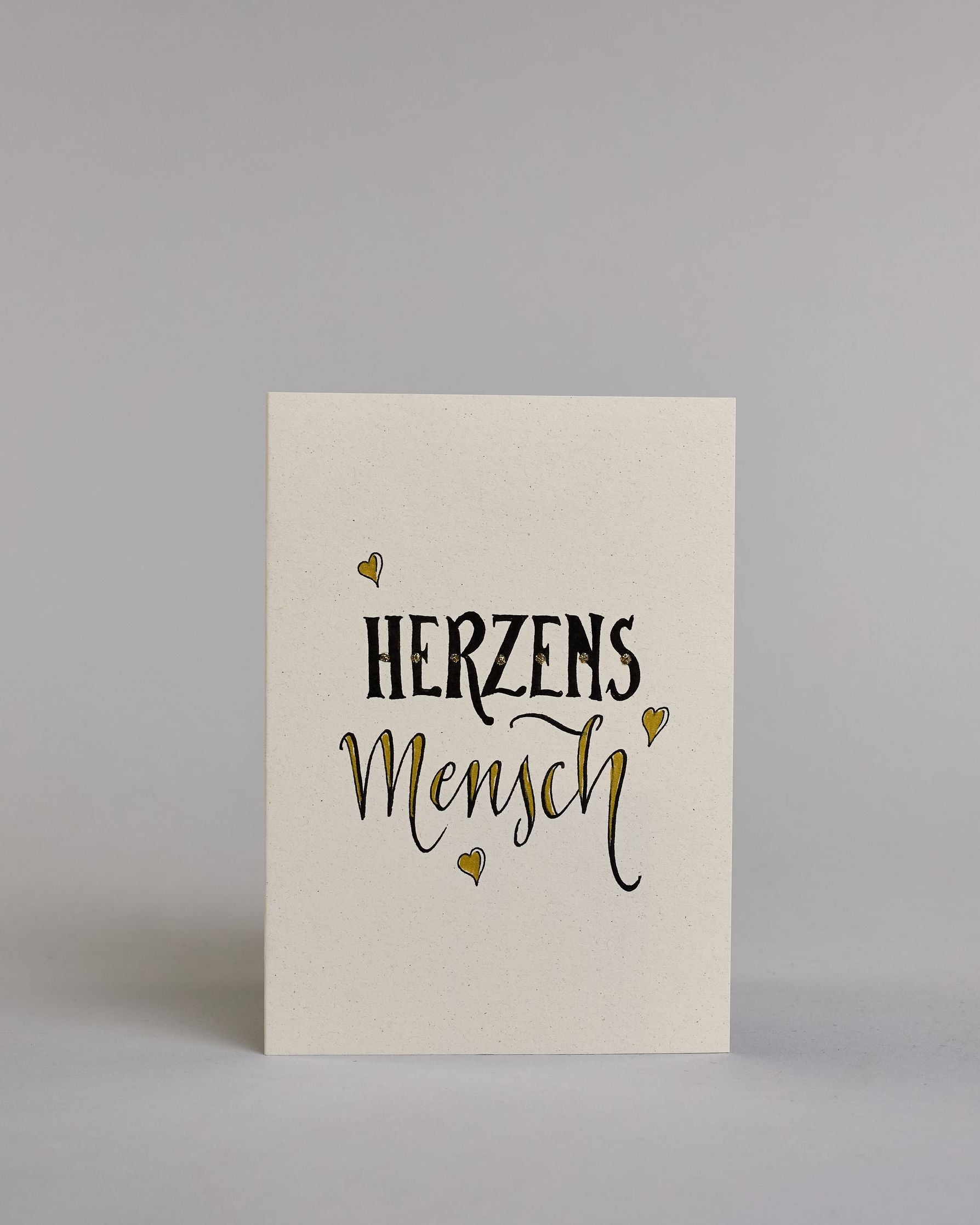 Klappkarte für Liebende mit schwarzer Kalligrafieaufschrift "Herzensmensch" mit drei gezeichneten schwebenden Herzen auf hellem Hintergrund. Der Text ist in zwei Zeilen aufgeteilt: "HERZENS" in schwarzer dicker, gerader Kalligrafieschrift, gold verziert und darunter "MENSCH" in verglitzerter Schreibschrift gestaltet, die Herzen befinden sich links über der ersten Zeile, rechts zwischen den beiden Zeilen und recht mittig unter der zweiten Zeile und sie sind gold bemalt. Dazu ein gold-weißes handgeschöpftes…
