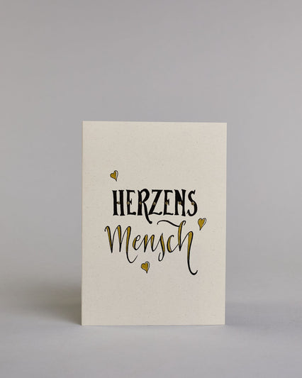 Klappkarte für Liebende mit schwarzer Kalligrafieaufschrift "Herzensmensch" mit drei gezeichneten schwebenden Herzen auf hellem Hintergrund. Der Text ist in zwei Zeilen aufgeteilt: "HERZENS" in schwarzer dicker, gerader Kalligrafieschrift, gold verziert und darunter "MENSCH" in verglitzerter Schreibschrift gestaltet, die Herzen befinden sich links über der ersten Zeile, rechts zwischen den beiden Zeilen und recht mittig unter der zweiten Zeile und sie sind gold bemalt. Dazu ein gold-weißes handgeschöpftes…