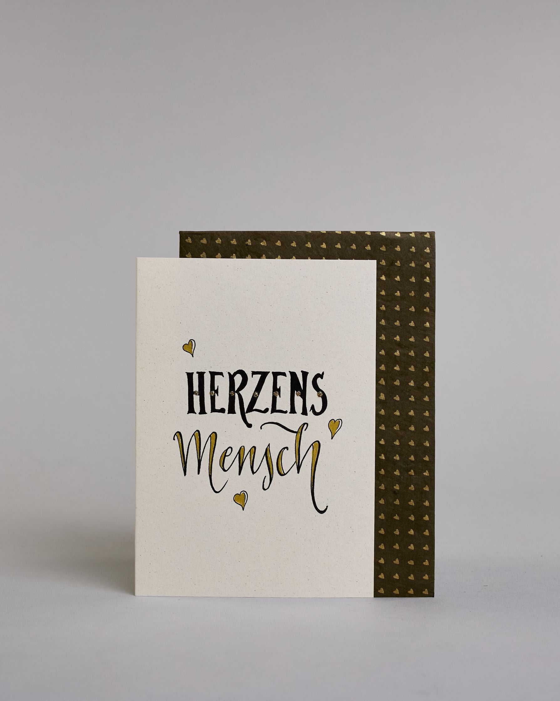 Klappkarte für Liebende mit schwarzer Kalligrafieaufschrift "Herzensmensch" mit drei gezeichneten schwebenden Herzen auf hellem Hintergrund. Der Text ist in zwei Zeilen aufgeteilt: "HERZENS" in schwarzer dicker, gerader Kalligrafieschrift, gold verziert und darunter "MENSCH" in verglitzerter Schreibschrift gestaltet, die Herzen befinden sich links über der ersten Zeile, rechts zwischen den beiden Zeilen und recht mittig unter der zweiten Zeile und sie sind gold bemalt. Dazu ein gold-weißes handgeschöpftes…