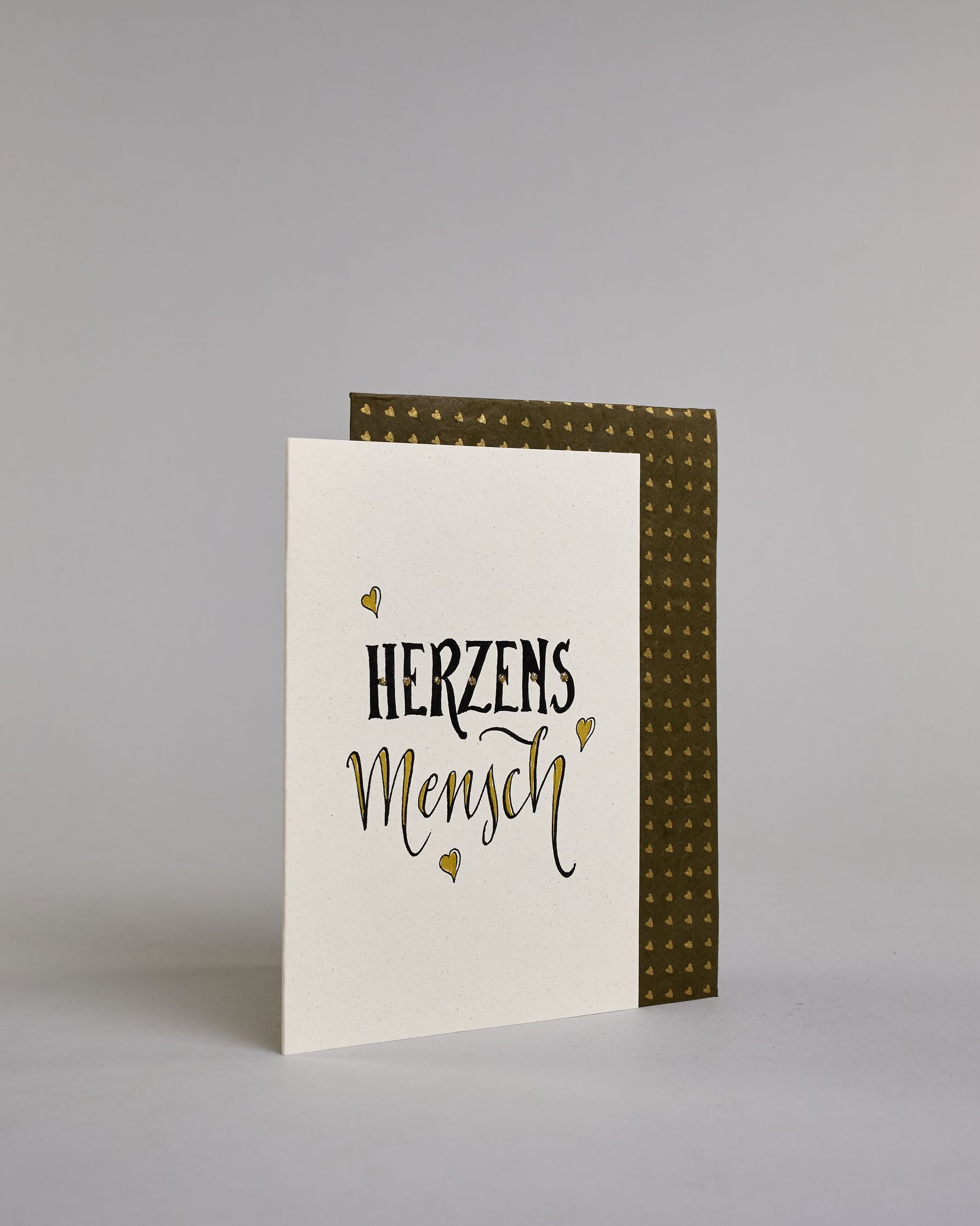 Klappkarte für Liebende mit schwarzer Kalligrafieaufschrift "Herzensmensch" mit drei gezeichneten schwebenden Herzen auf hellem Hintergrund. Der Text ist in zwei Zeilen aufgeteilt: "HERZENS" in schwarzer dicker, gerader Kalligrafieschrift, gold verziert und darunter "MENSCH" in verglitzerter Schreibschrift gestaltet, die Herzen befinden sich links über der ersten Zeile, rechts zwischen den beiden Zeilen und recht mittig unter der zweiten Zeile und sie sind gold bemalt. Dazu ein gold-weißes handgeschöpftes…