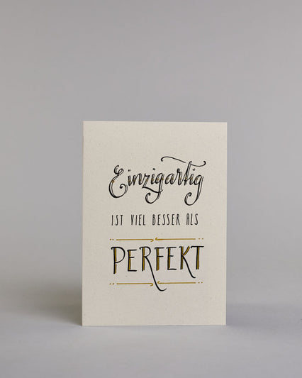 Klappkarte für Liebende mit schwarzer Kalligrafieaufschrift "Einzigartig ist viel besser als perfekt" auf hellem Hintergrund. Der Text ist in folgende drei Zeilen aufgeteilt: "Einzigartig" in prunkvoller Schreibschrift und Goldverzierung rundherum glitzernde Punkte, darunter "IST VIEL BESSER ALS" in dünner gerader Kalligrafieschrift, darunter "PERFEKT" in verspielter gerader Kalligrafieschift welche zwischen zwei horizontalen Linien die in der mitte mit Schnörkseln unterbrochen sind. Dazu ein grün-goldenes…