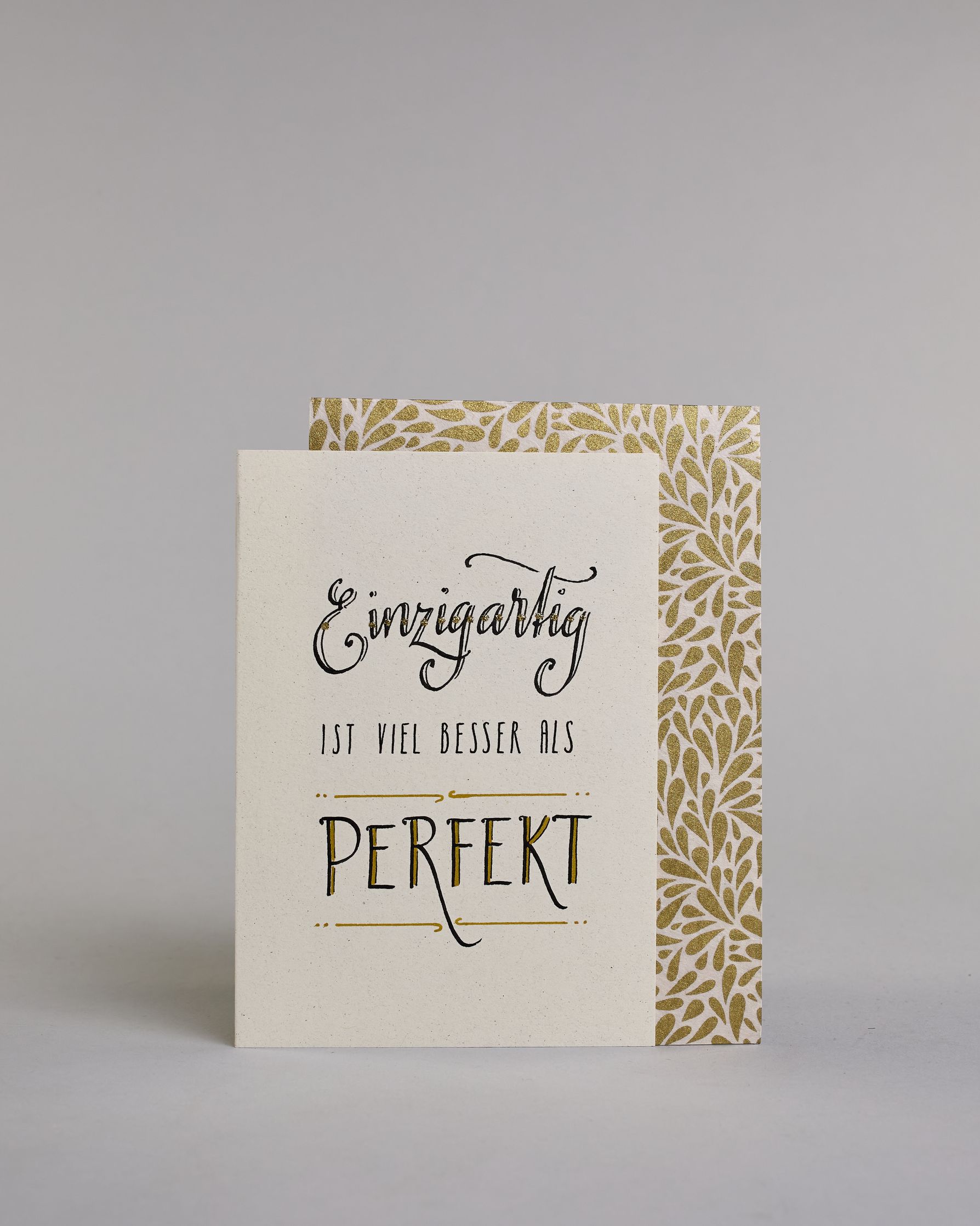 Klappkarte für Liebende mit schwarzer Kalligrafieaufschrift "Einzigartig ist viel besser als perfekt" auf hellem Hintergrund. Der Text ist in folgende drei Zeilen aufgeteilt: "Einzigartig" in prunkvoller Schreibschrift und Goldverzierung rundherum glitzernde Punkte, darunter "IST VIEL BESSER ALS" in dünner gerader Kalligrafieschrift, darunter "PERFEKT" in verspielter gerader Kalligrafieschift welche zwischen zwei horizontalen Linien die in der mitte mit Schnörkseln unterbrochen sind. Dazu ein grün-goldenes…