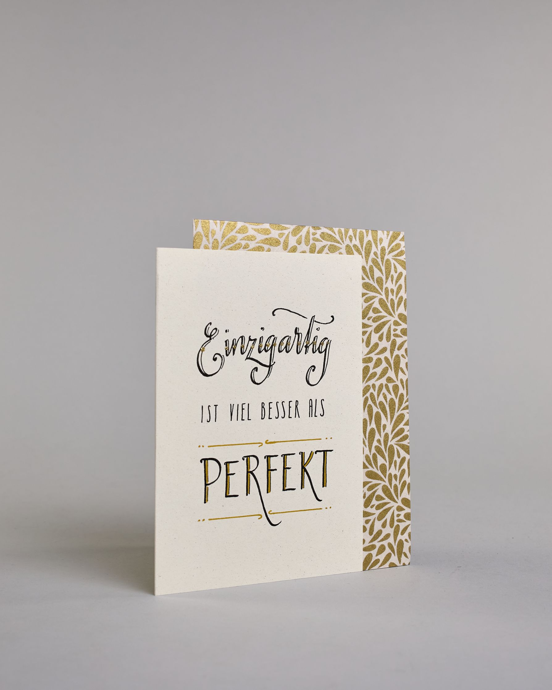 Klappkarte für Liebende mit schwarzer Kalligrafieaufschrift "Einzigartig ist viel besser als perfekt" auf hellem Hintergrund. Der Text ist in folgende drei Zeilen aufgeteilt: "Einzigartig" in prunkvoller Schreibschrift und Goldverzierung rundherum glitzernde Punkte, darunter "IST VIEL BESSER ALS" in dünner gerader Kalligrafieschrift, darunter "PERFEKT" in verspielter gerader Kalligrafieschift welche zwischen zwei horizontalen Linien die in der mitte mit Schnörkseln unterbrochen sind. Dazu ein grün-goldenes…