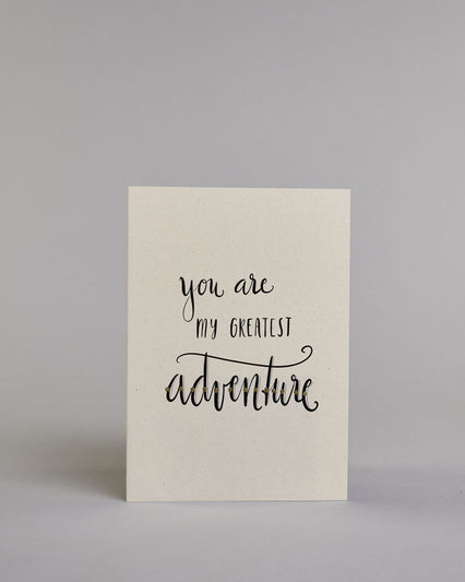 Klappkarte für Liebende in Englisch mit schwarzer Kalligrafieaufschrift "You are my greatest adventure" auf hellem Hintergrund. Der Text ist in folgende drei Zeilen aufgeteilt: "you are" in geschwungener Schreibschrift und glitzernden Punkten in der Mitte der Buchstaben als Verzierung, darunter "MY GREATEST" in verspielter gerader Kalligrafieschrift, darunter "adventure" in der gleichen Schreibschift wie in der ersten Zeile, hier ist der Text jedoch komplett gold verziert. Dazu ein gold-weißes handgeschöpf…