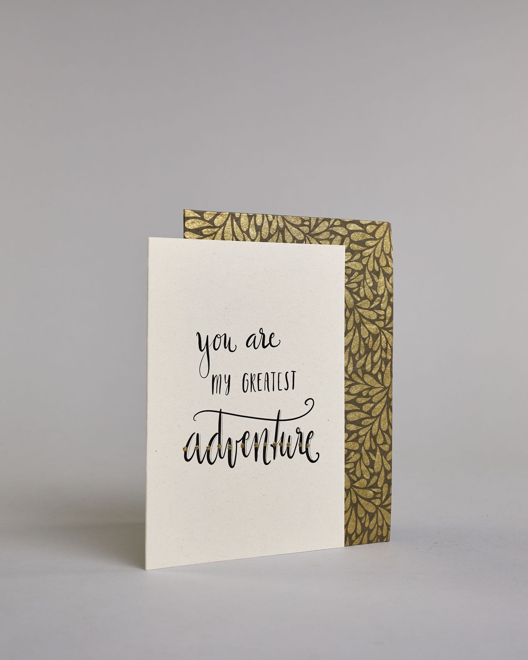 Klappkarte für Liebende in Englisch mit schwarzer Kalligrafieaufschrift "You are my greatest adventure" auf hellem Hintergrund. Der Text ist in folgende drei Zeilen aufgeteilt: "you are" in geschwungener Schreibschrift und glitzernden Punkten in der Mitte der Buchstaben als Verzierung, darunter "MY GREATEST" in verspielter gerader Kalligrafieschrift, darunter "adventure" in der gleichen Schreibschift wie in der ersten Zeile, hier ist der Text jedoch komplett gold verziert. Dazu ein gold-weißes handgeschöpf…