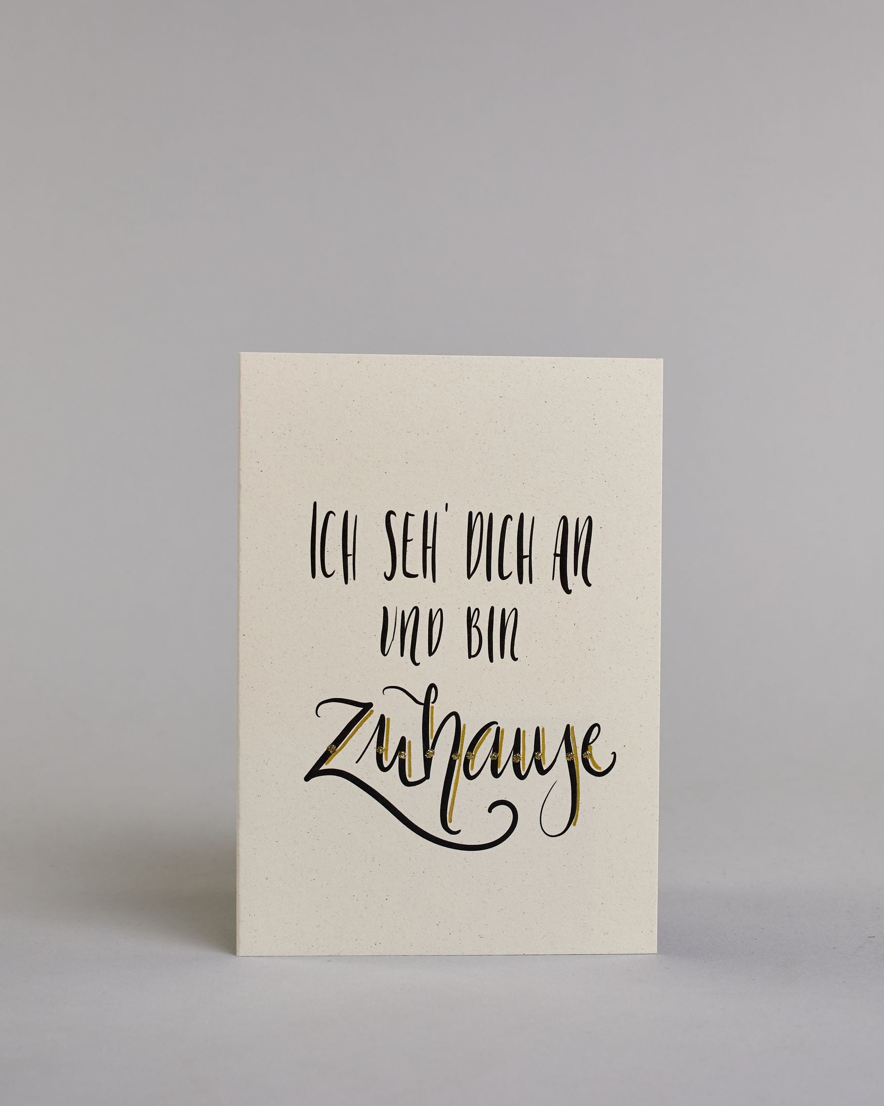 Klappkarte für Liebende mit schwarzer Kalligrafieaufschrift "Ich seh' dich an und bin Zuhause" auf hellem Hintergrund. Der Text ist in folgende drei Zeilen aufgeteilt: "ICH SEH' DICH AN", darunter "UND BIN" beide Zeilen sind in einer verspielten geraden Kalligrafieschrift gestaltet, darunter "Zuhause" in verspielter geschwungener Schreibschrift. Dazu ein handgeschöpftes Kuvert. #1